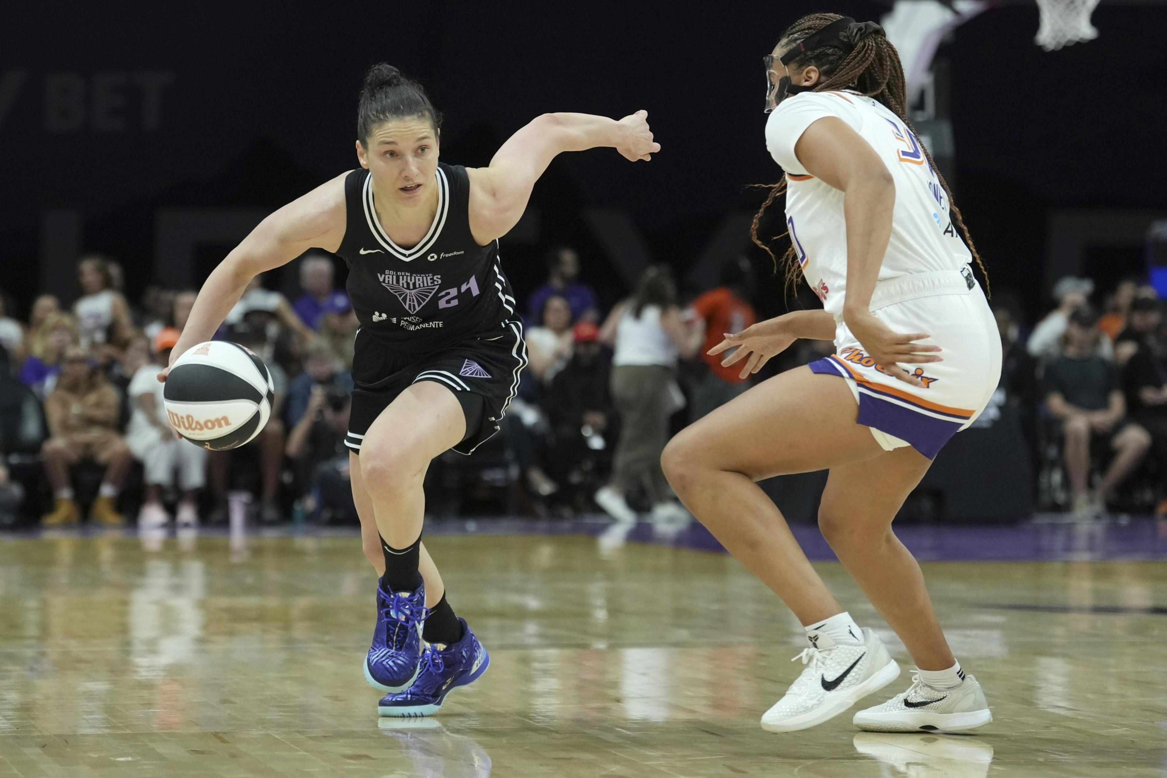 A basketball player in a black jersey dribbles while being guarded by a player in white. The court is indoors, with spectators in the background.