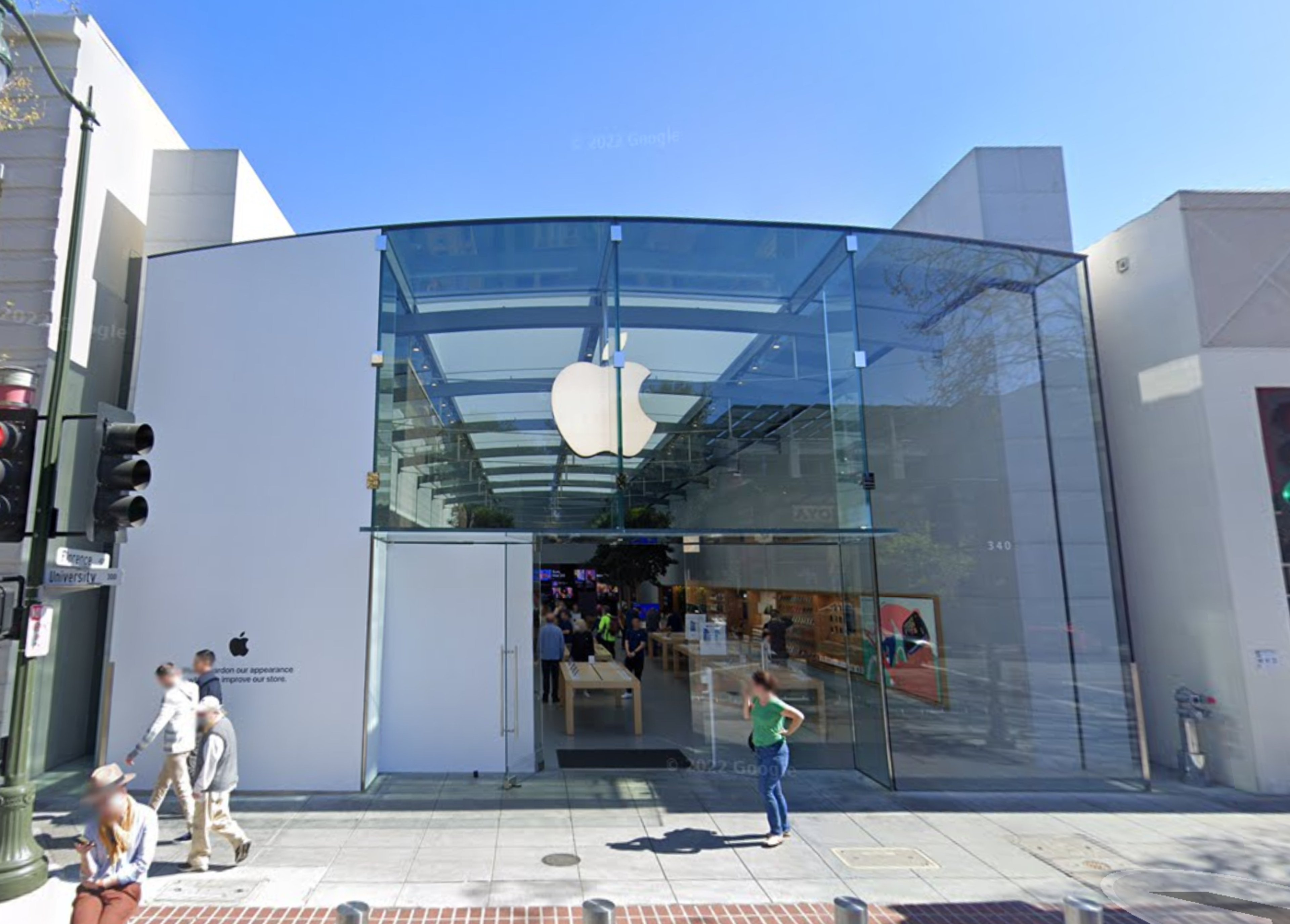 A modern Apple Store with a large glass facade and the Apple logo above the entrance, people walking on the sidewalk, and clear sunny weather.