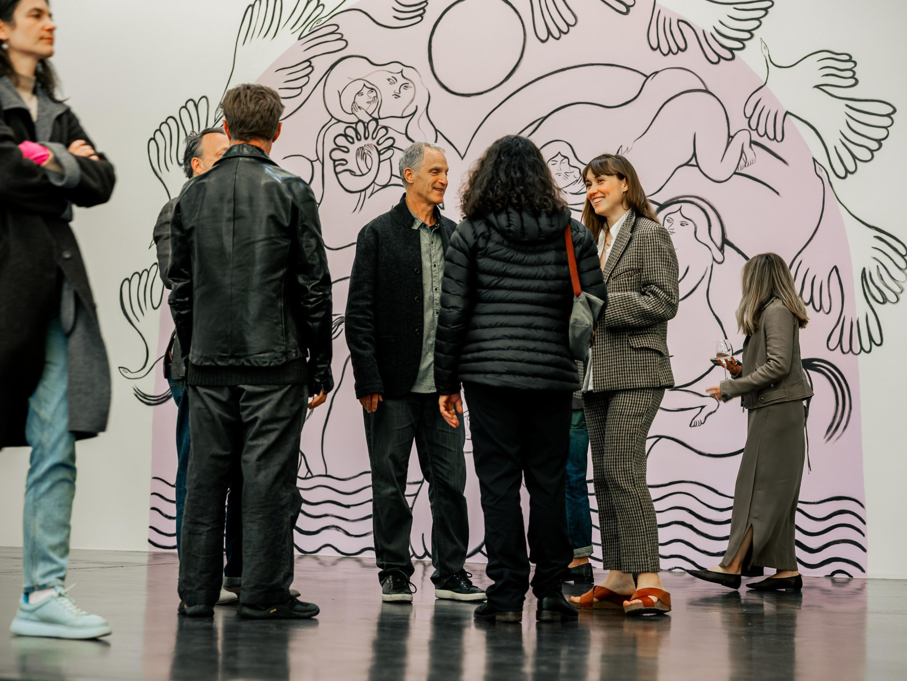 A group of people are standing in an art gallery, talking in front of a pastel artwork depicting abstract human and bird forms.