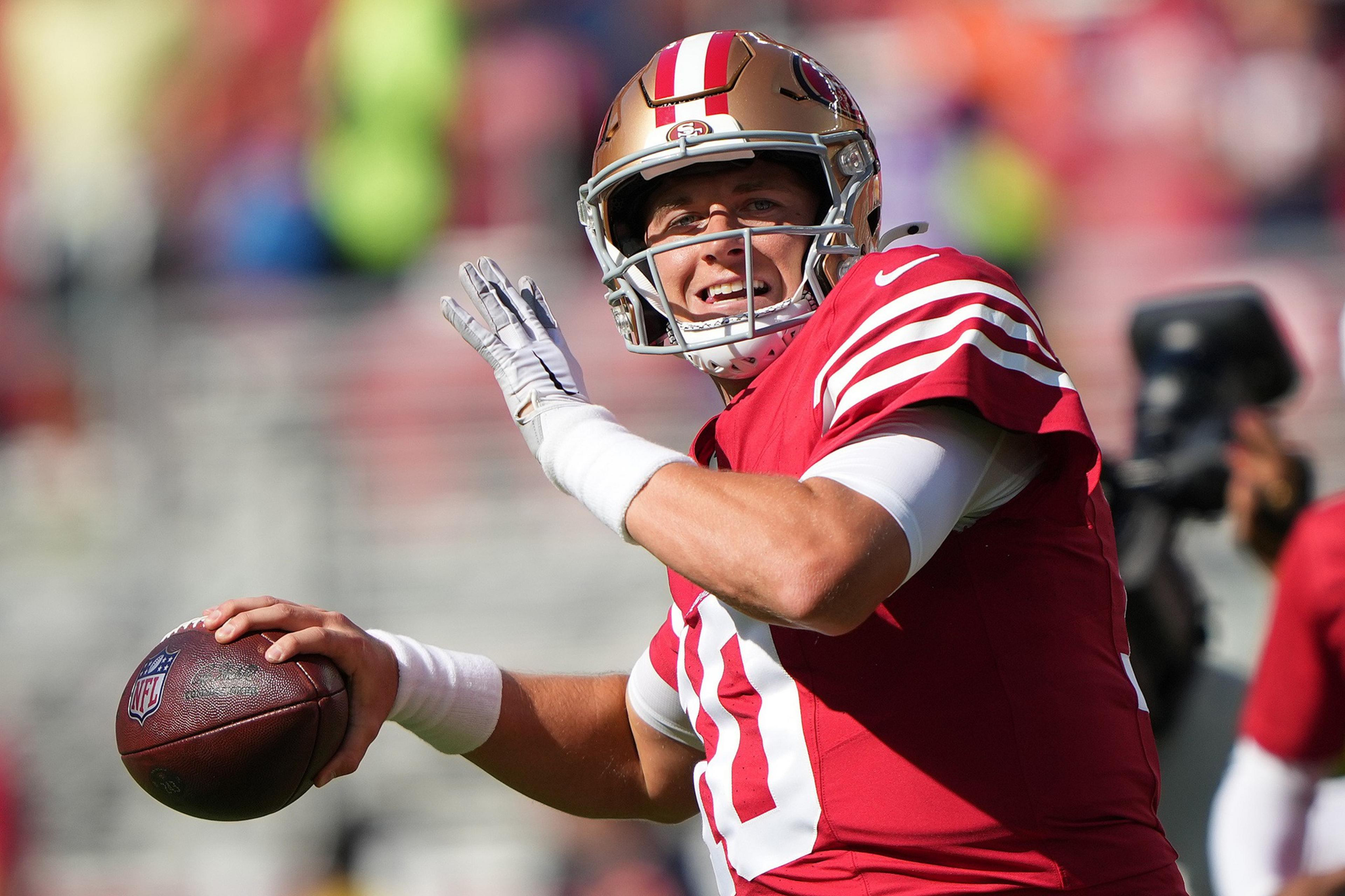A football player in a red and gold uniform prepares to throw a football during a sunny game, with a focused yet smiling expression.
