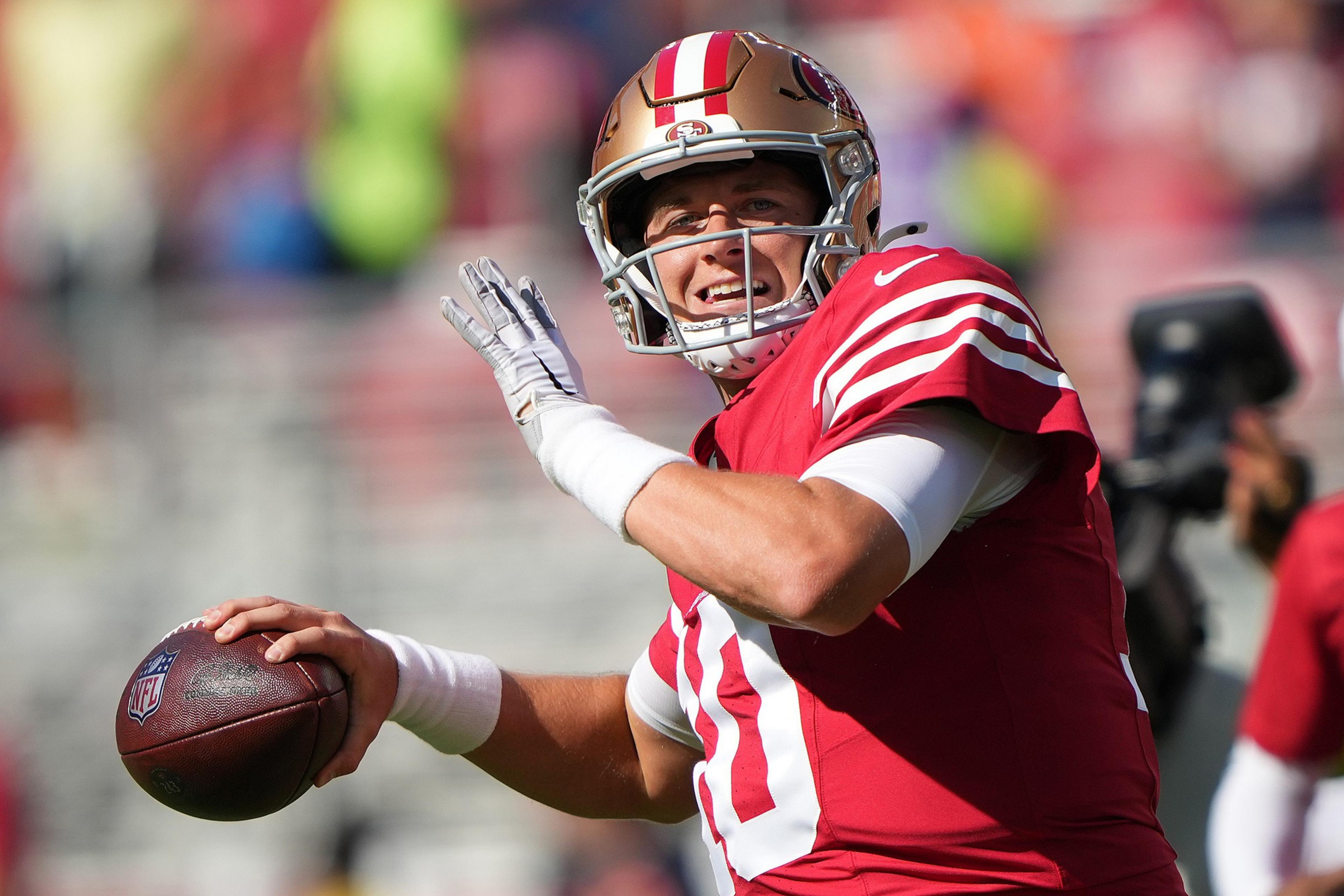 A football player in a red and gold uniform prepares to throw a football during a sunny game, with a focused yet smiling expression.