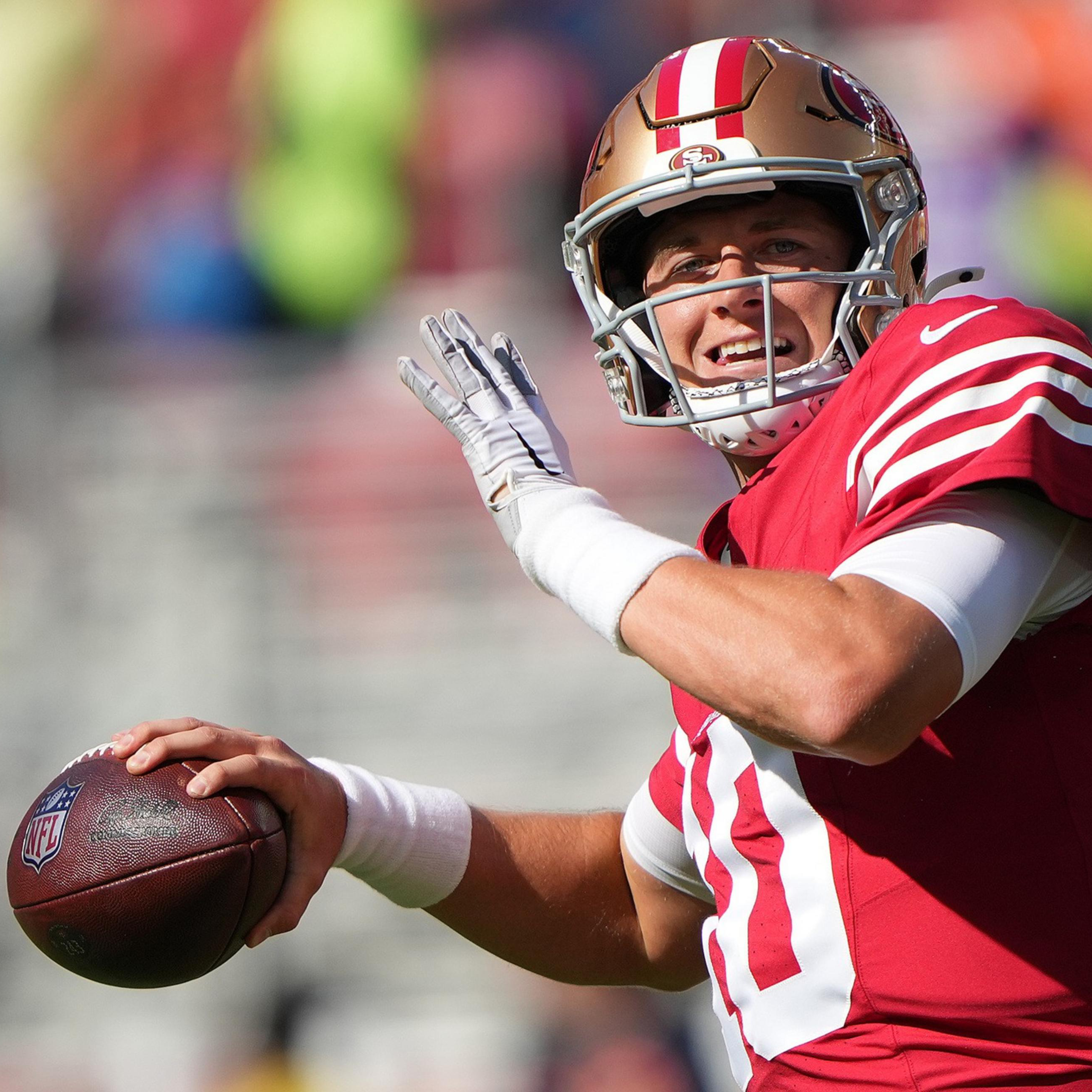 A football player in a red and gold uniform prepares to throw a football during a sunny game, with a focused yet smiling expression.