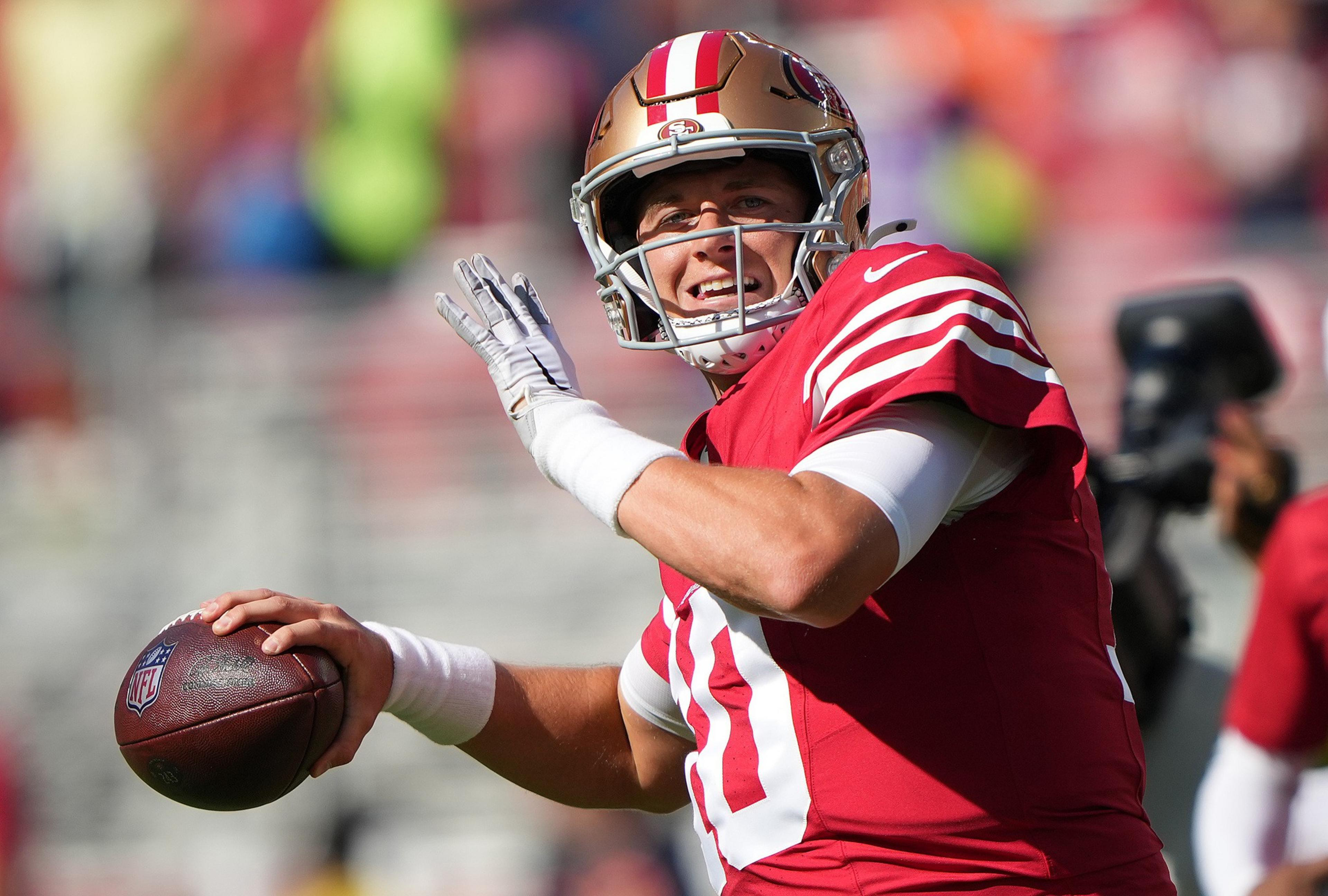 A football player in a red and gold uniform prepares to throw a football during a sunny game, with a focused yet smiling expression.