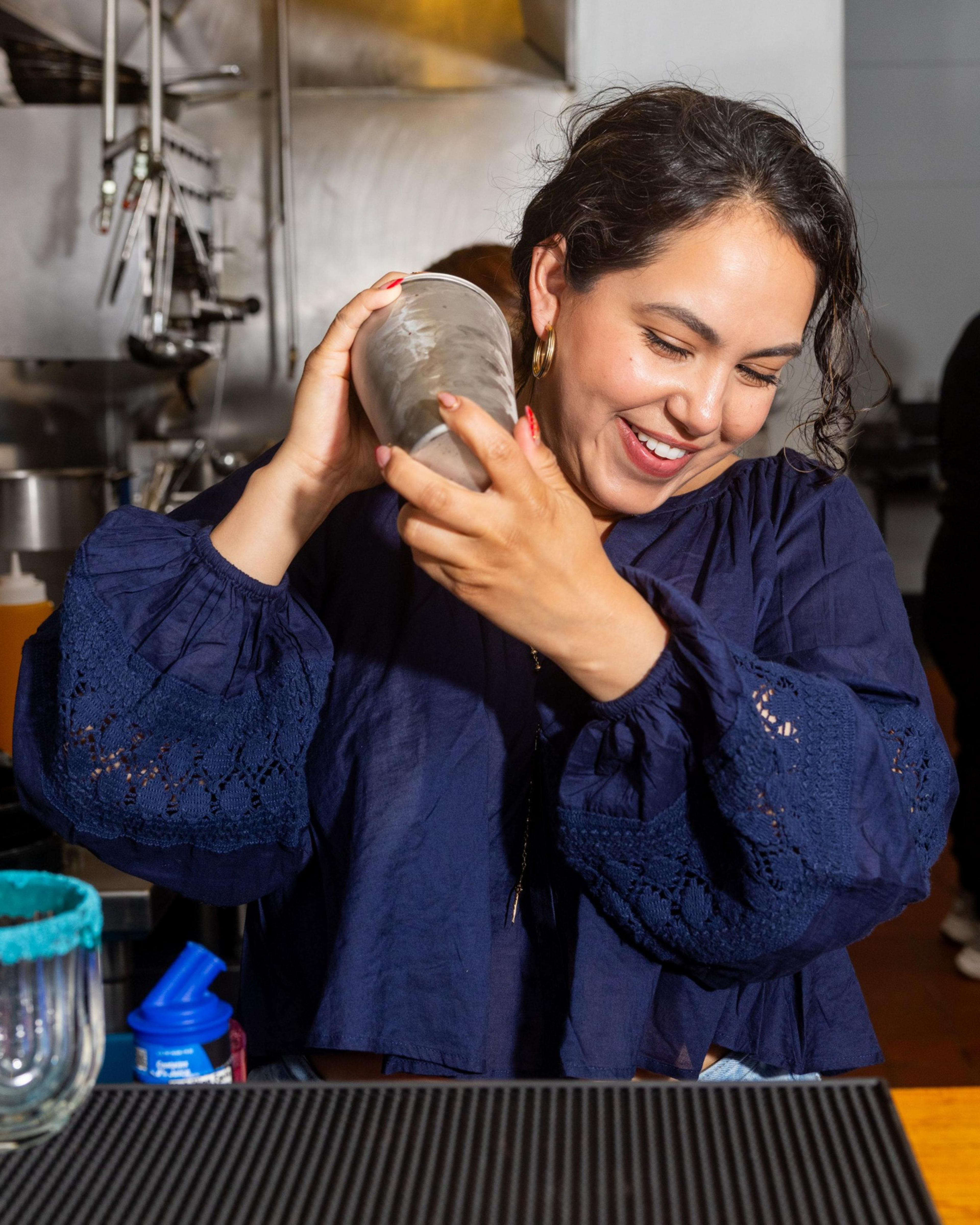 A woman with dark hair and a navy blouse is smiling while shaking a cocktail shaker in a kitchen. A glass with a blue rim is in the foreground.