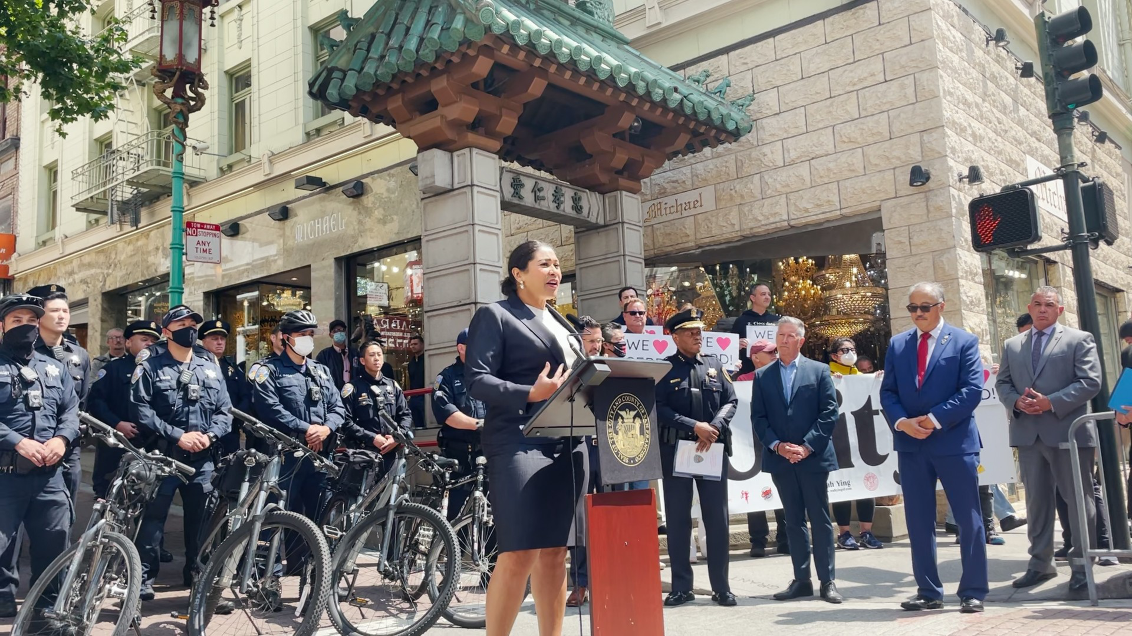 A woman speaks at a podium with a city seal, surrounded by police officers, officials in suits, and people holding "We ❤️" signs near a decorative building entrance.