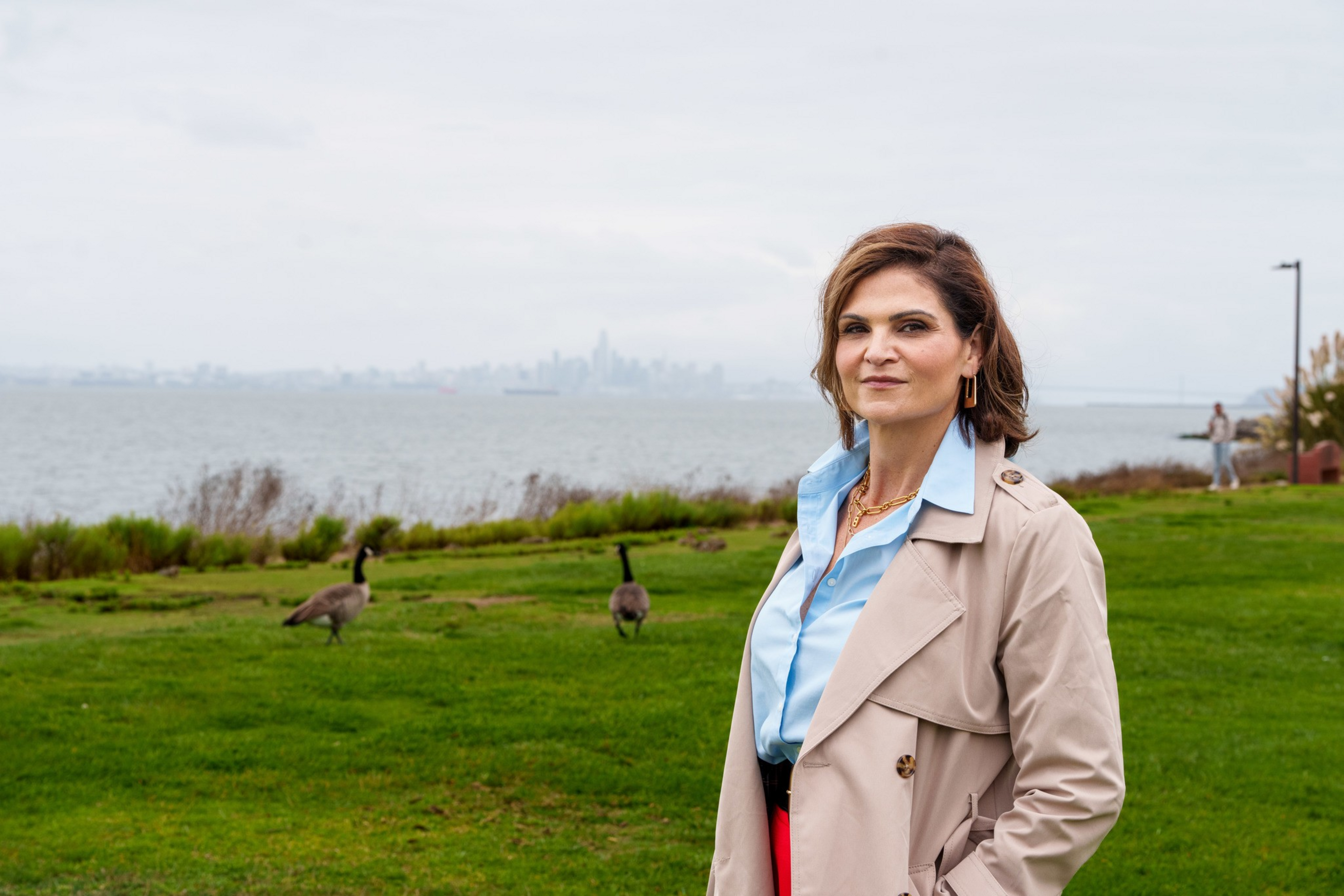 A woman in a beige trench coat and blue shirt stands on a grassy area by the water with two geese behind her and a city skyline in the distance.