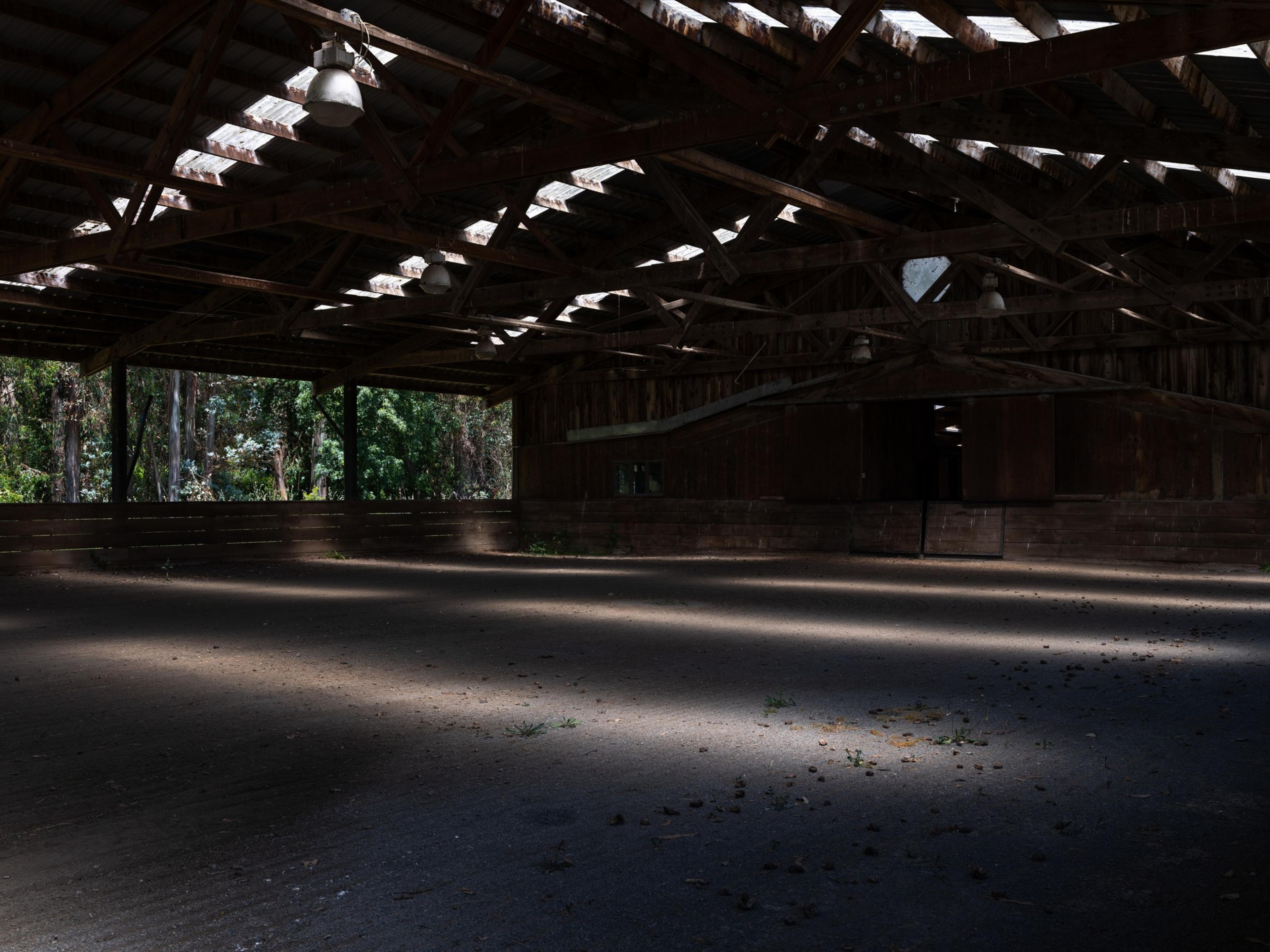 The image shows a dimly lit, empty wooden structure with a high ceiling and beams. Sunlight streams in through gaps, creating patterns on the dusty floor.