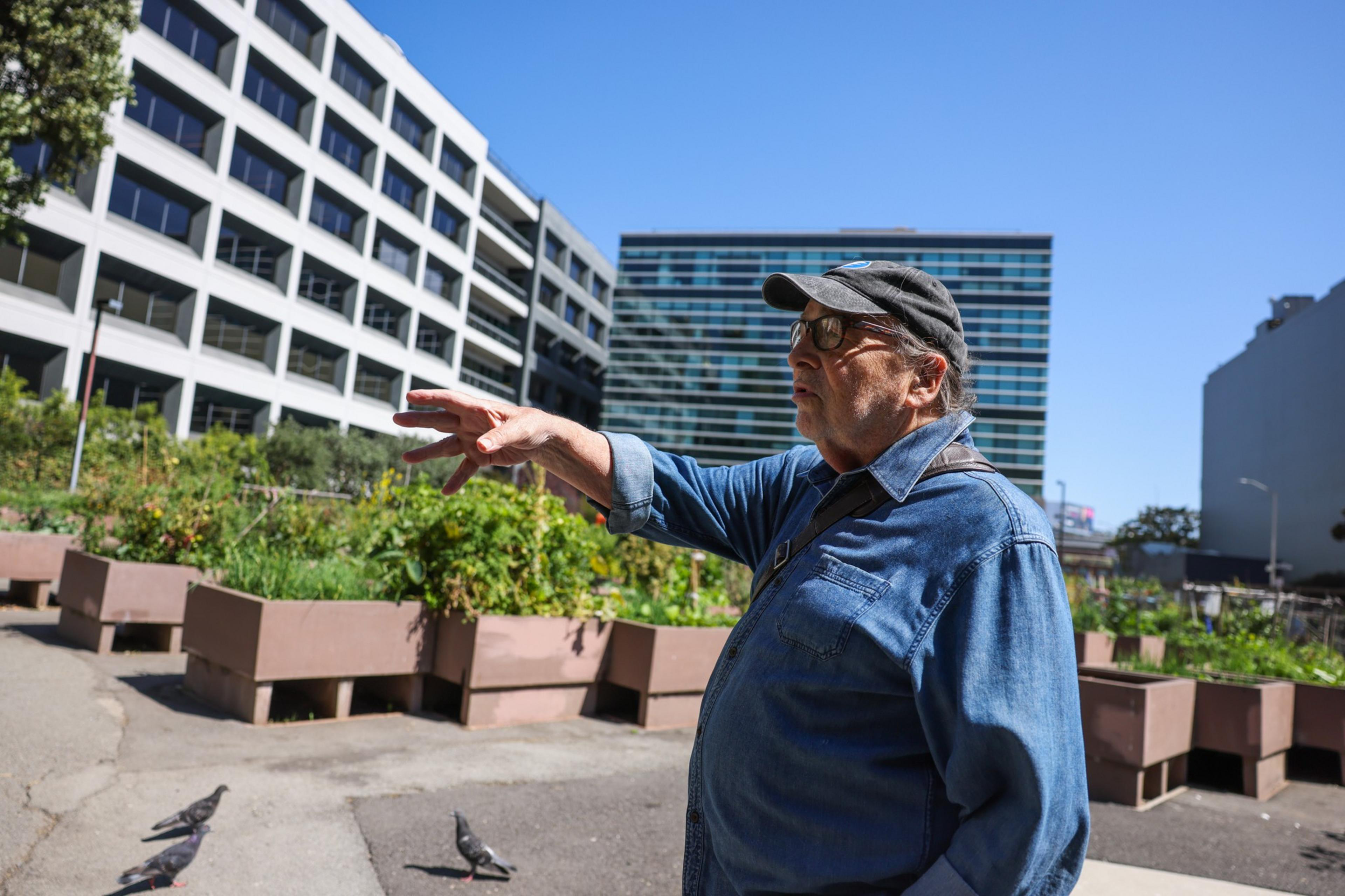 An older man wearing a denim shirt and cap gestures with his hand near urban garden beds, surrounded by modern office buildings on a sunny day.