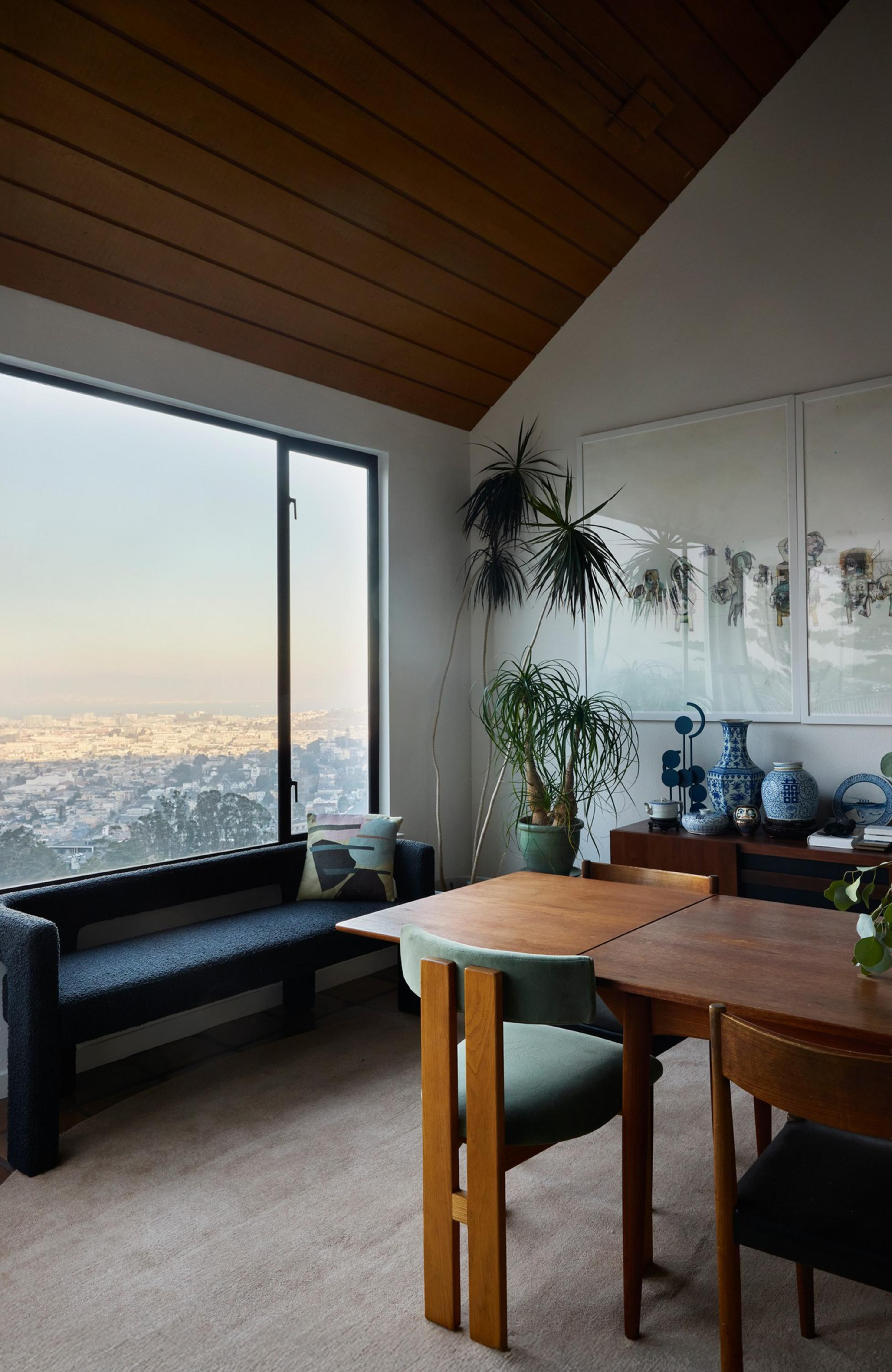 A cozy room with a large window overlooking a city, featuring a wooden table with chairs, a dark bench with pillows, tall plants, and decorative vases on a sideboard.