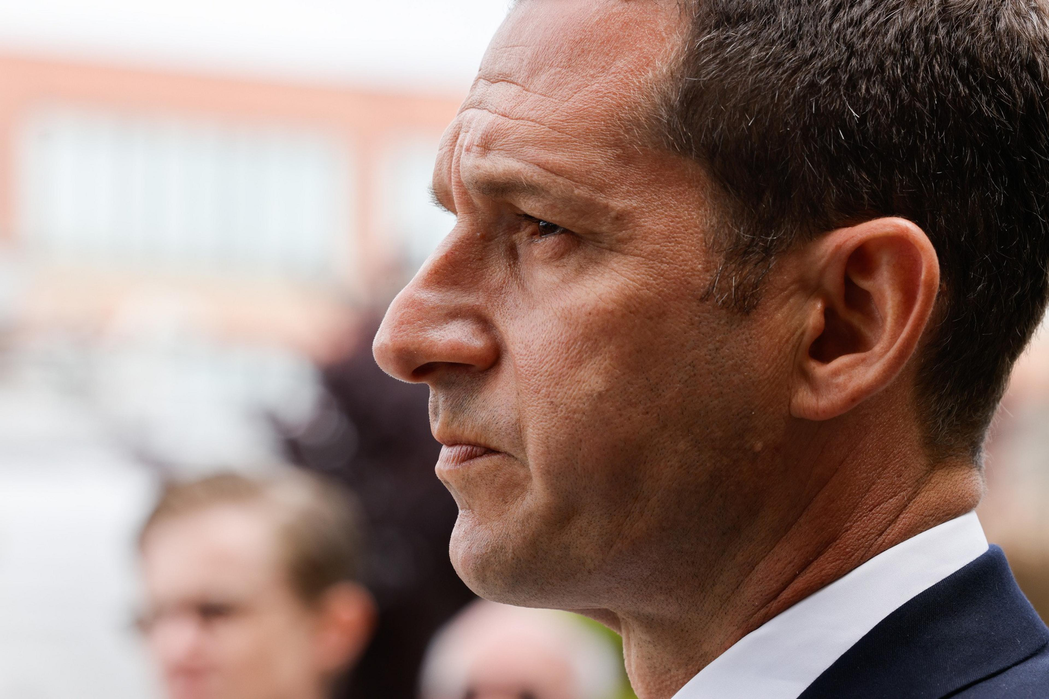 A man in a suit looks intently ahead with a serious expression, while blurred figures and buildings appear in the background.
