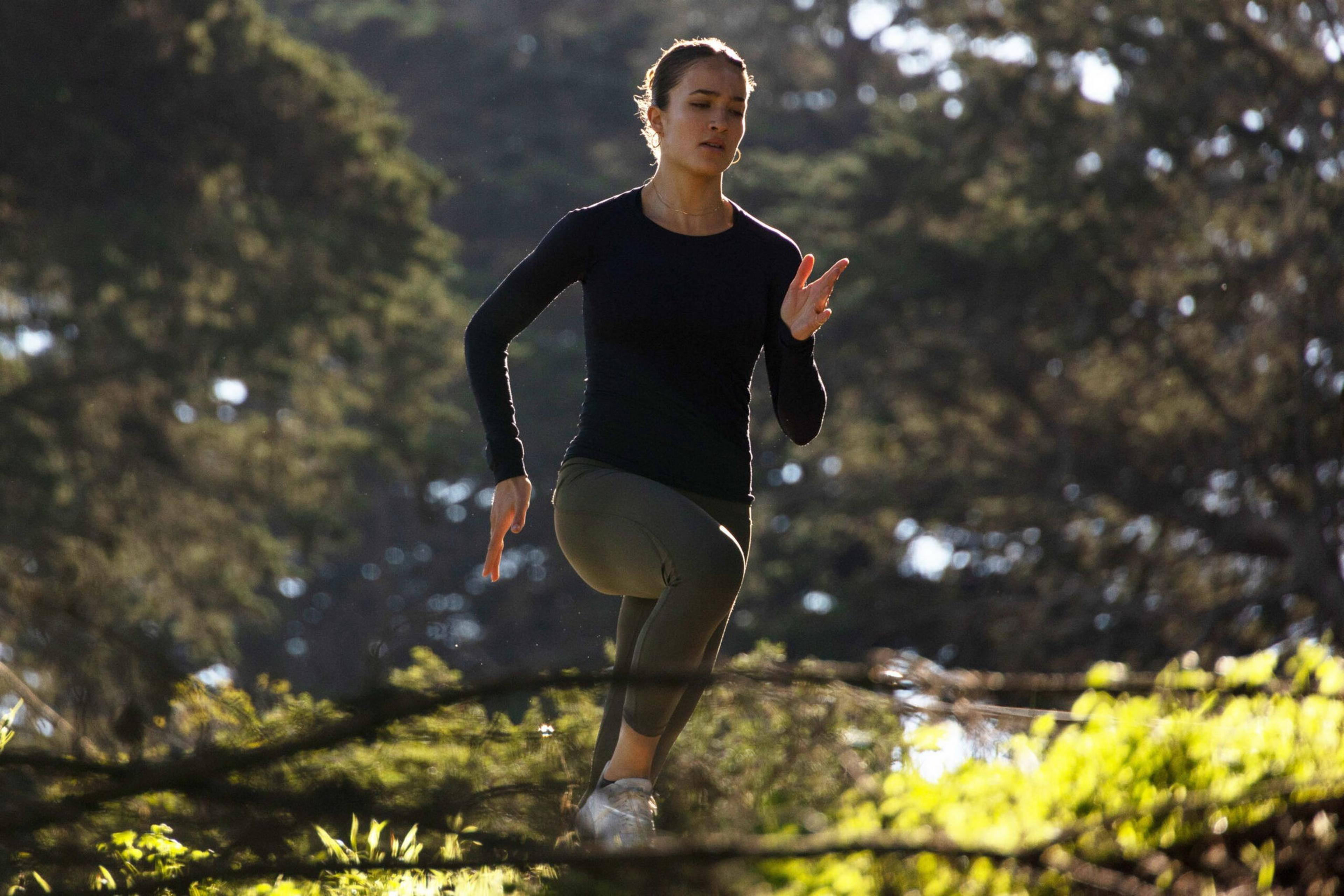 A woman in a black long-sleeve shirt and green leggings is jogging outdoors on a sunny day, surrounded by trees and greenery.