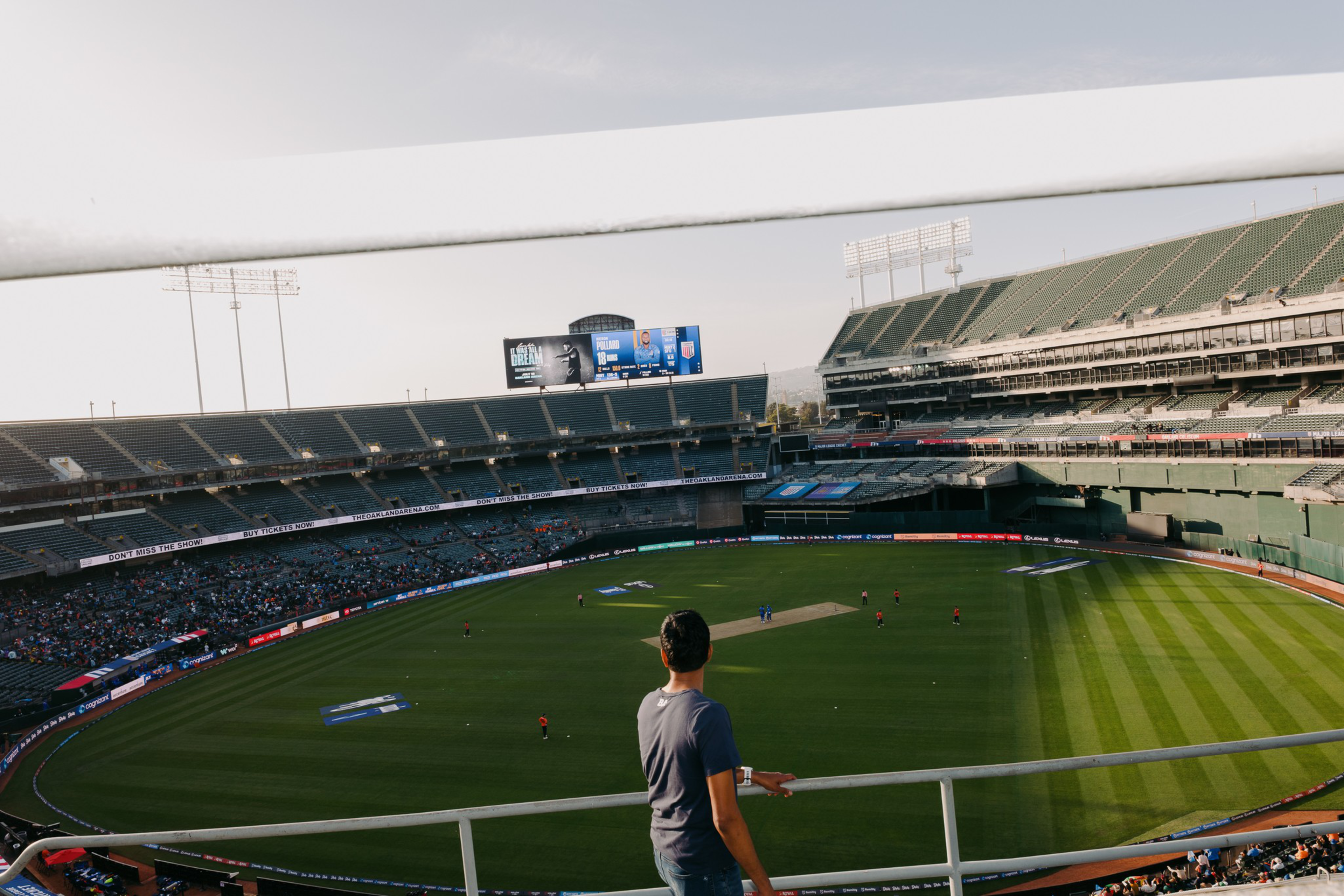 A man is watching a cricket match in a large stadium with sparse spectators. The field is green, and scoreboards are visible in the distance.