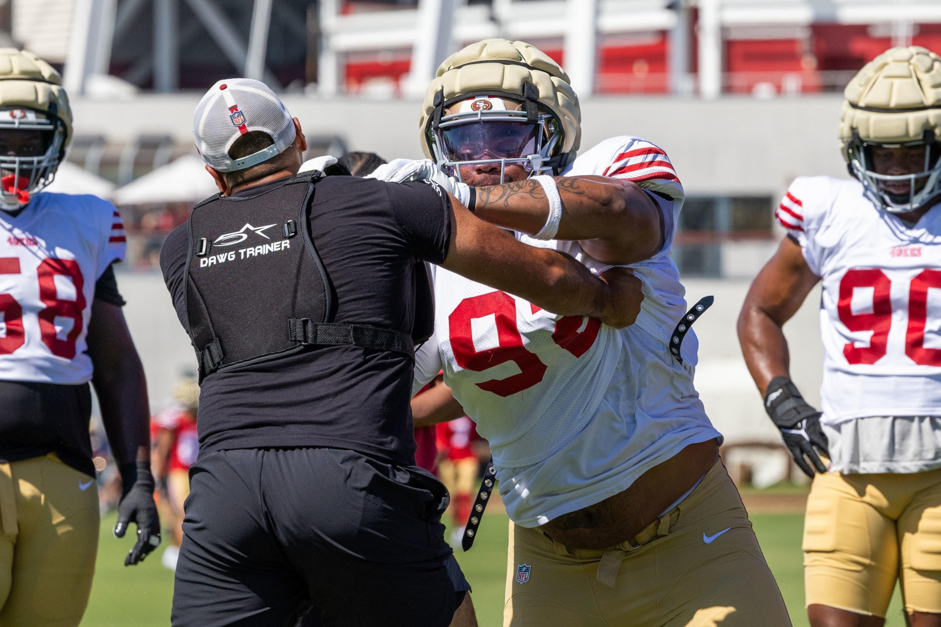 A football player wearing number 98 practices blocking against a trainer wearing black gear labeled "Dawg Trainer," with teammates in the background.