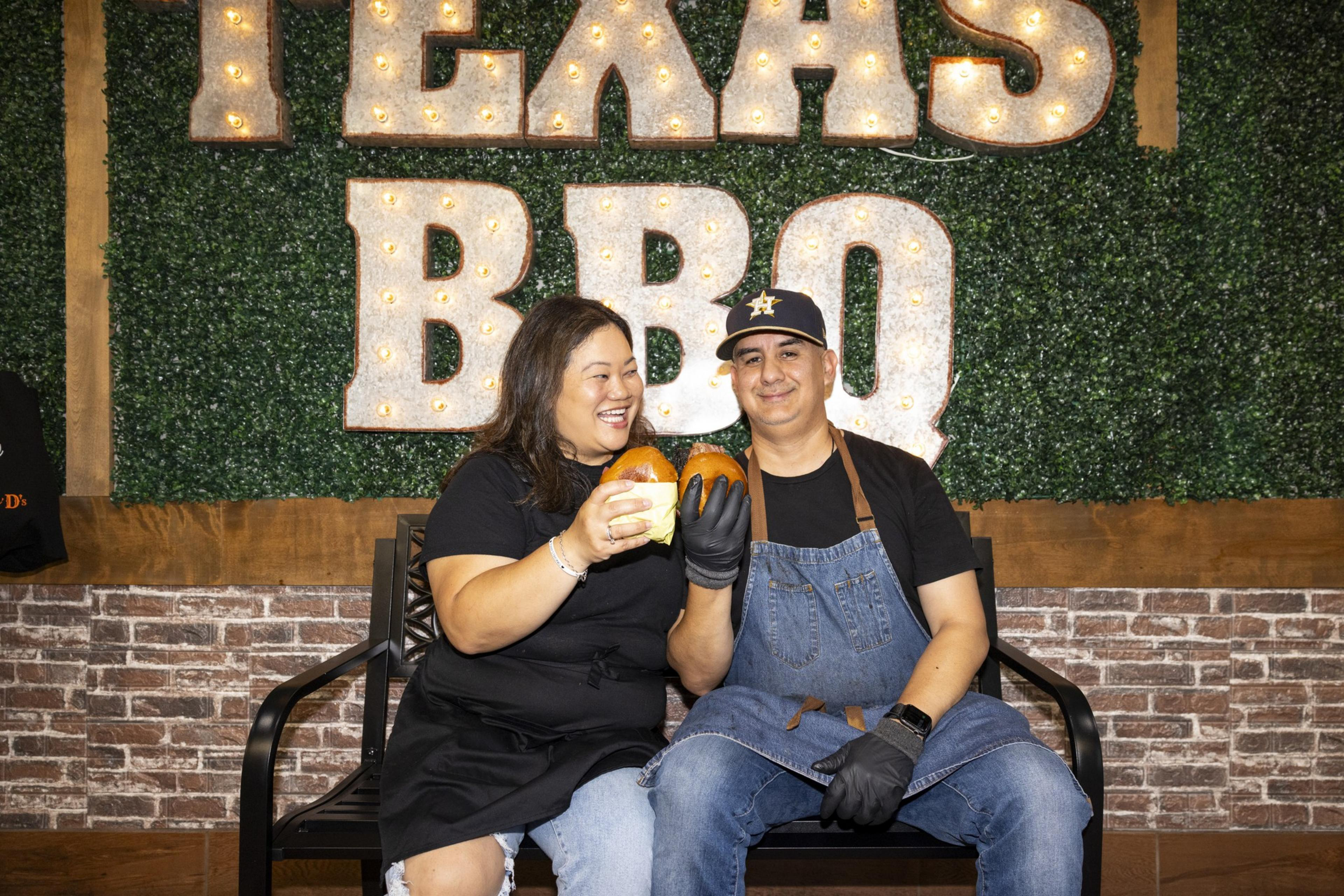 A smiling man and woman sit on a bench holding two burgers in front of a wall with illuminated "Texas BBQ" sign, with a rustic brick and plant background.