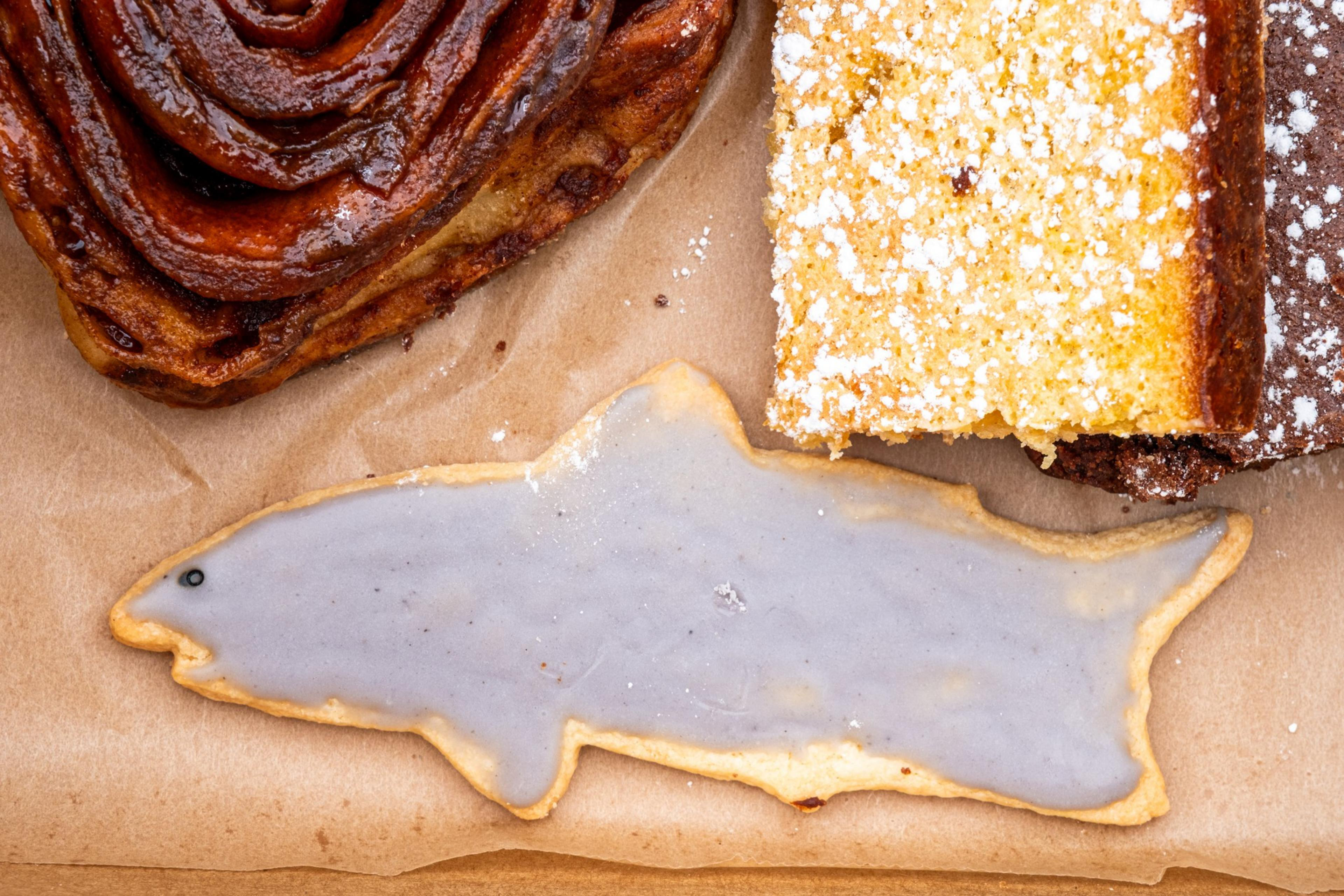 A shark-shaped cookie with light purple icing sits next to a glossy, spiral pastry and a powdered sugar-dusted slice of cake on brown paper.