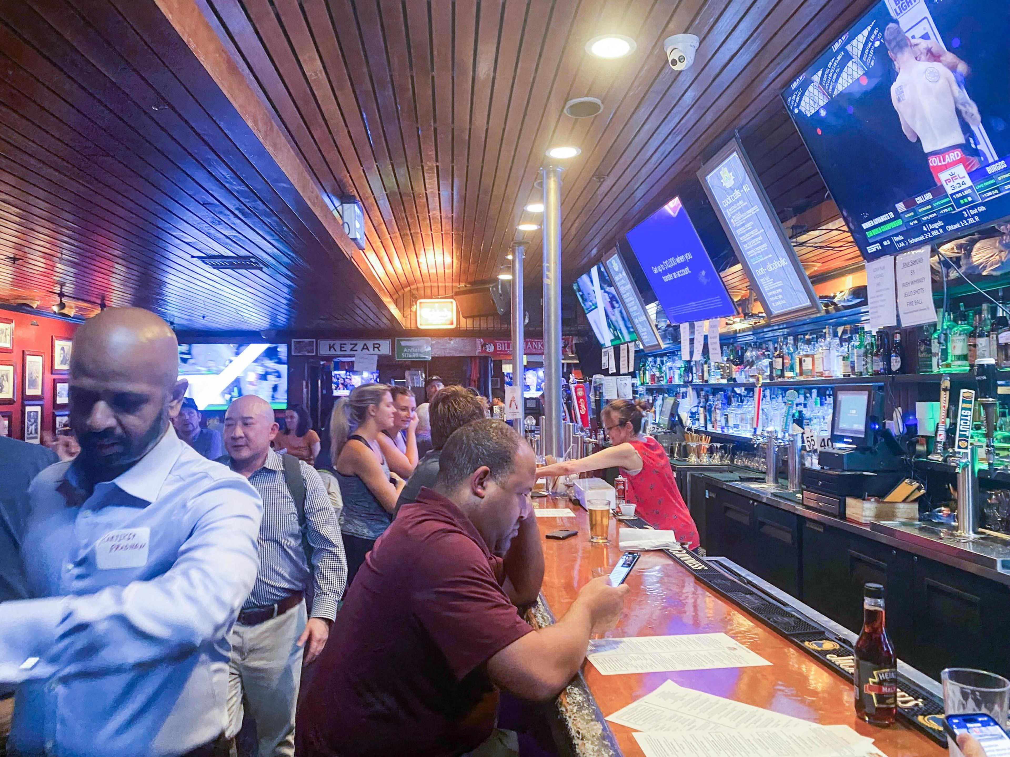 It's a busy bar with people socializing and drinking. TVs are mounted above the bar showing sports. A bartender serves drinks to patrons seated at the counter.