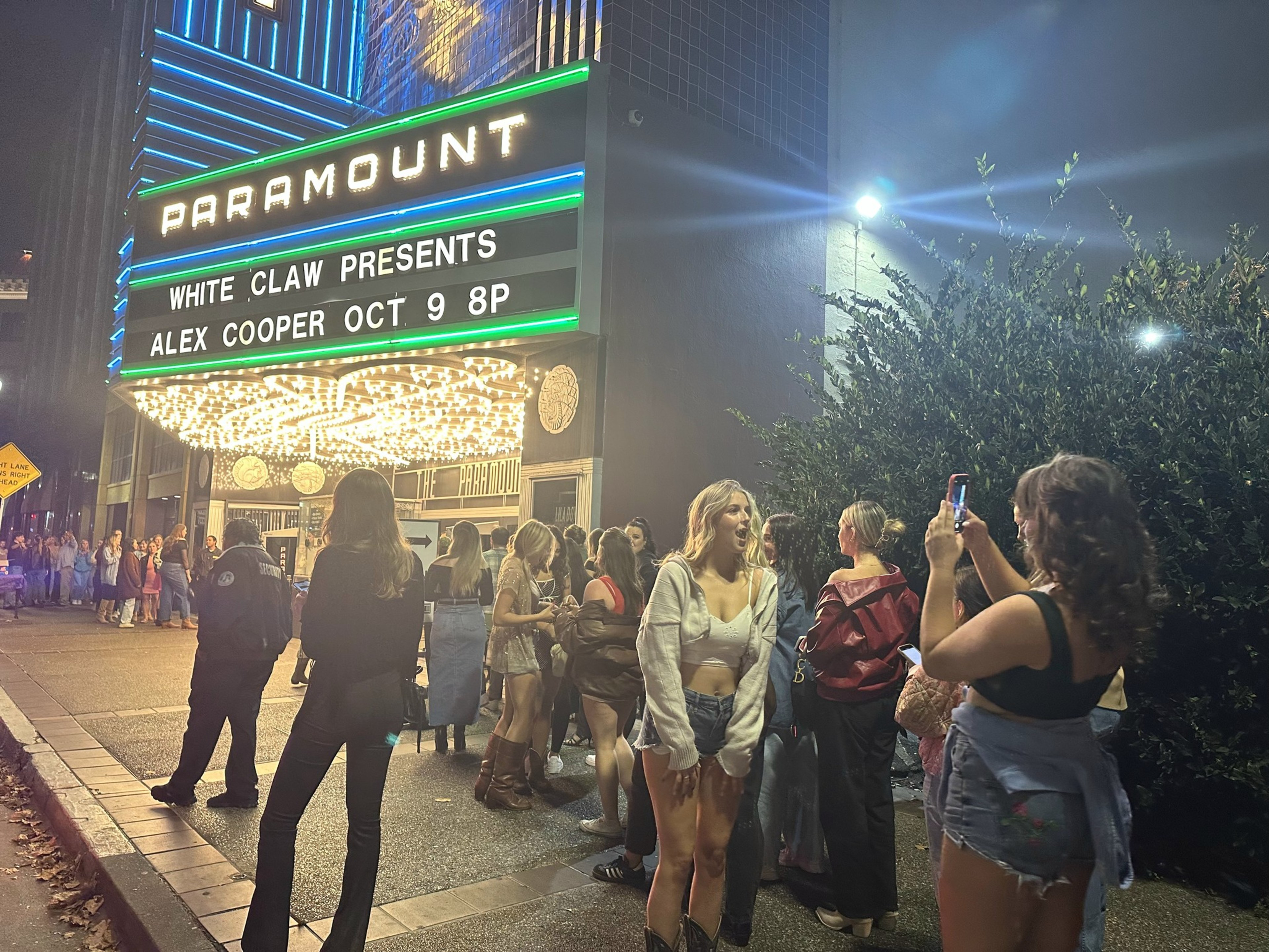 Under a neon marquee reading "Paramount" a long line of women stand waiting to get in as one woman in jean shorts and a white bralette poses for a photo.