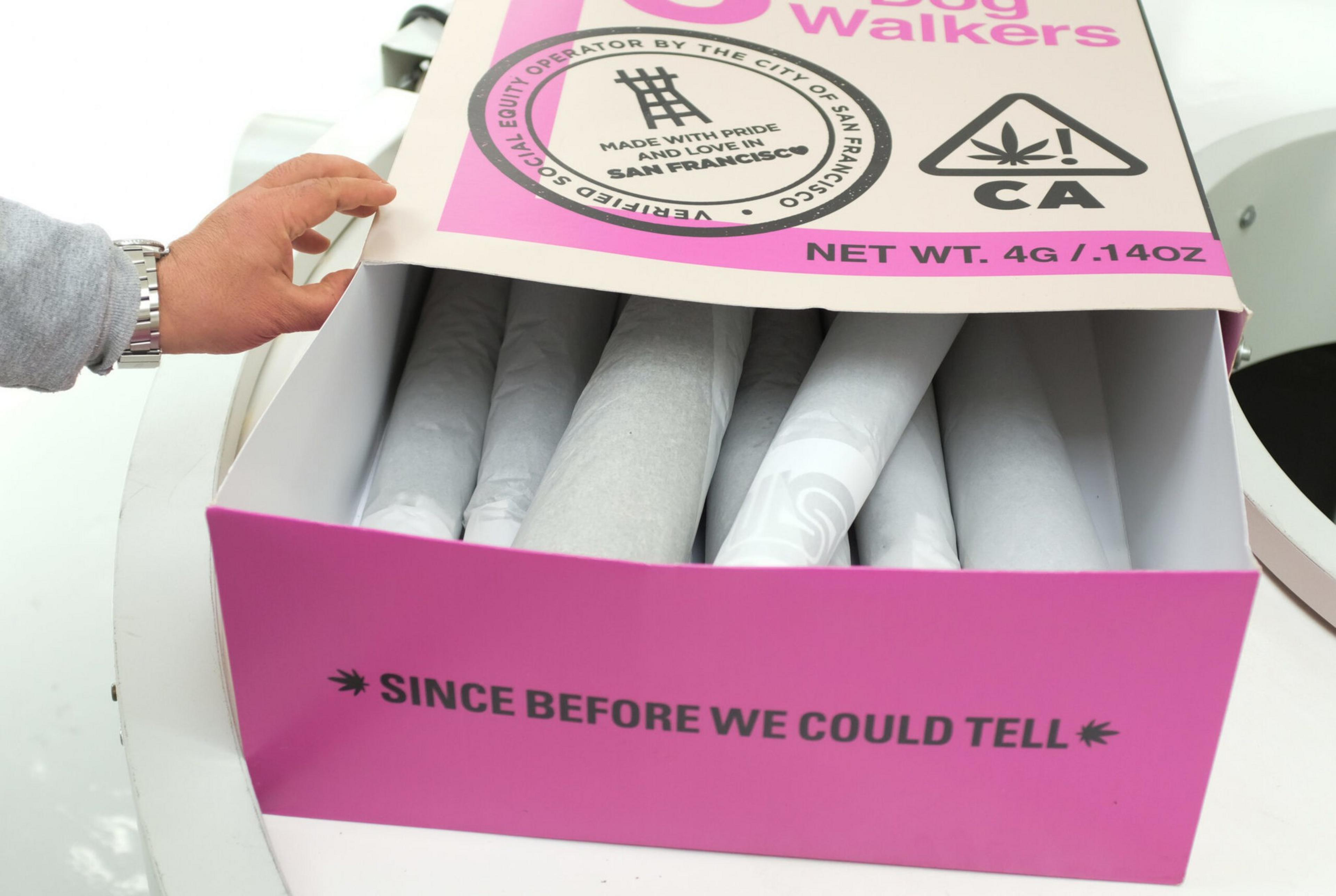 A hand lifts the lid of a pink box labeled "Since Before We Could Tell," revealing six white individually wrapped cylindrical items inside.