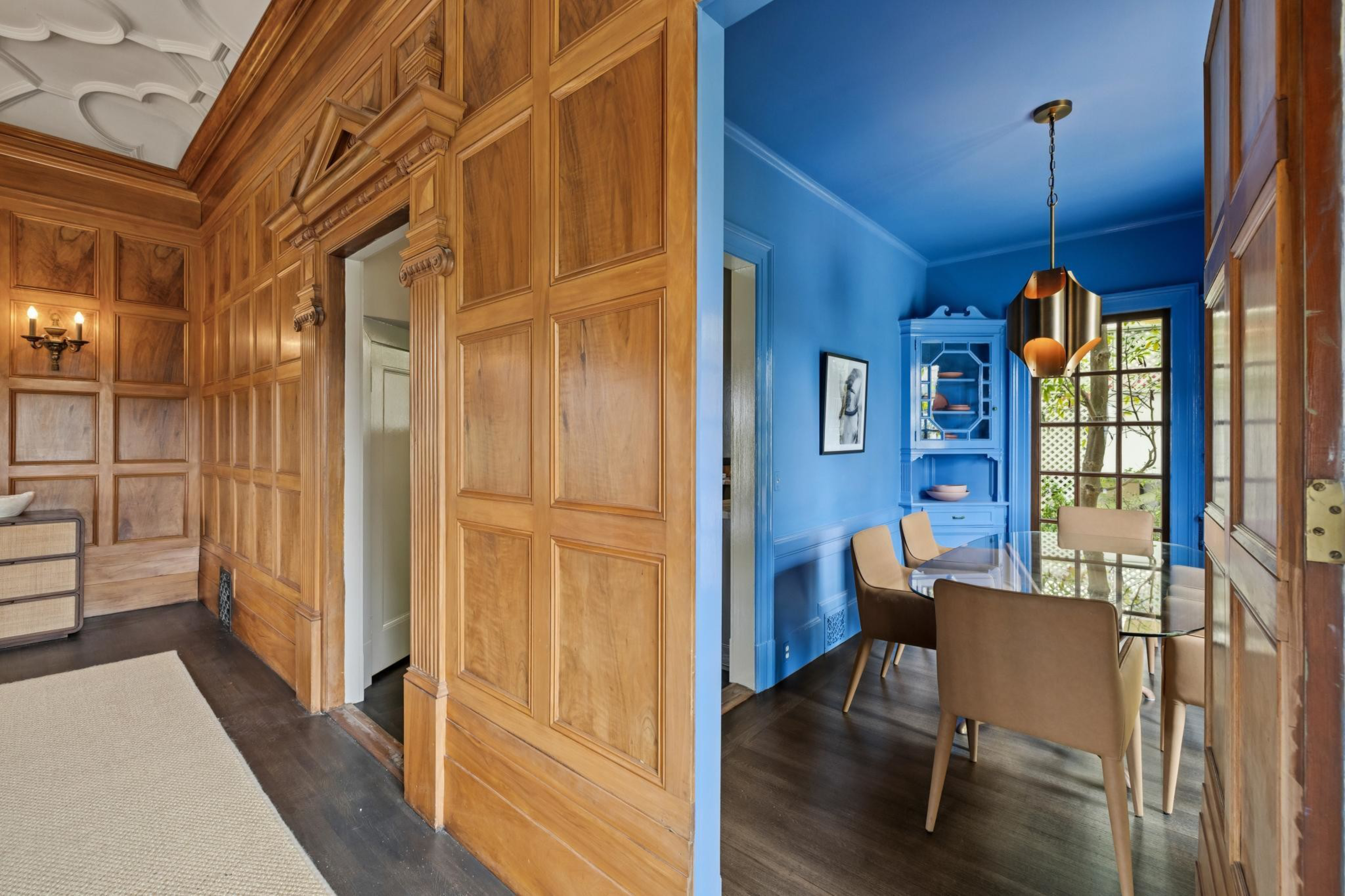 A hallway with detailed wood paneling leads into a vivid blue dining room featuring a glass table, beige chairs, a blue cabinet, and a modern pendant light.