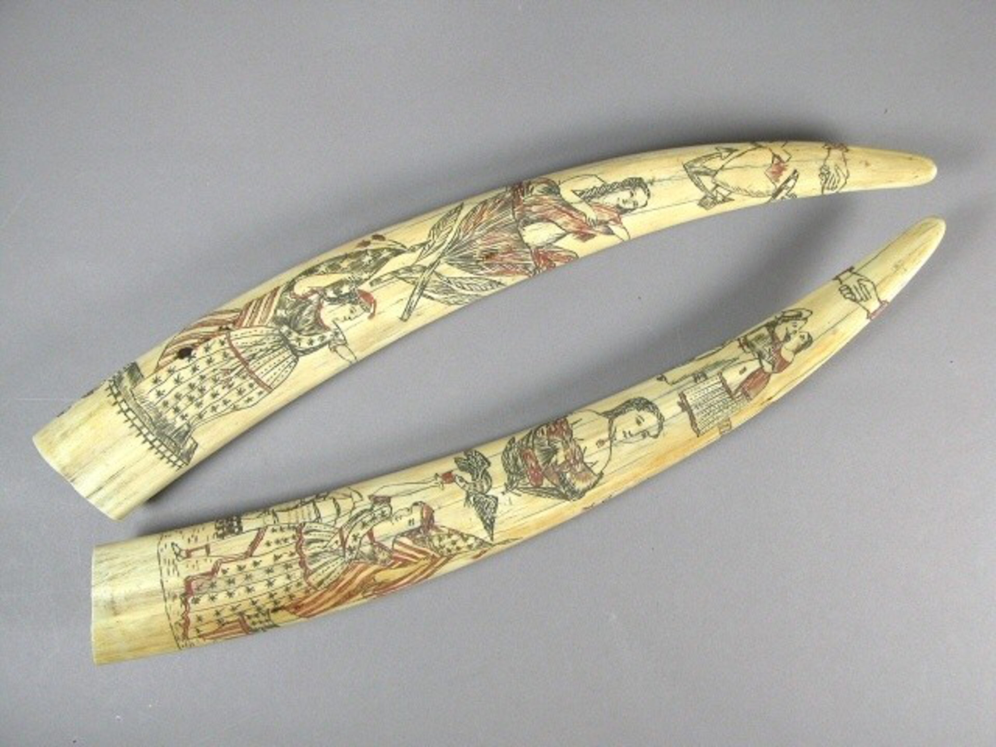 Two carved ivory whale teeth feature detailed scrimshaw artwork depicting colonial-era figures, including men and women in period clothing.