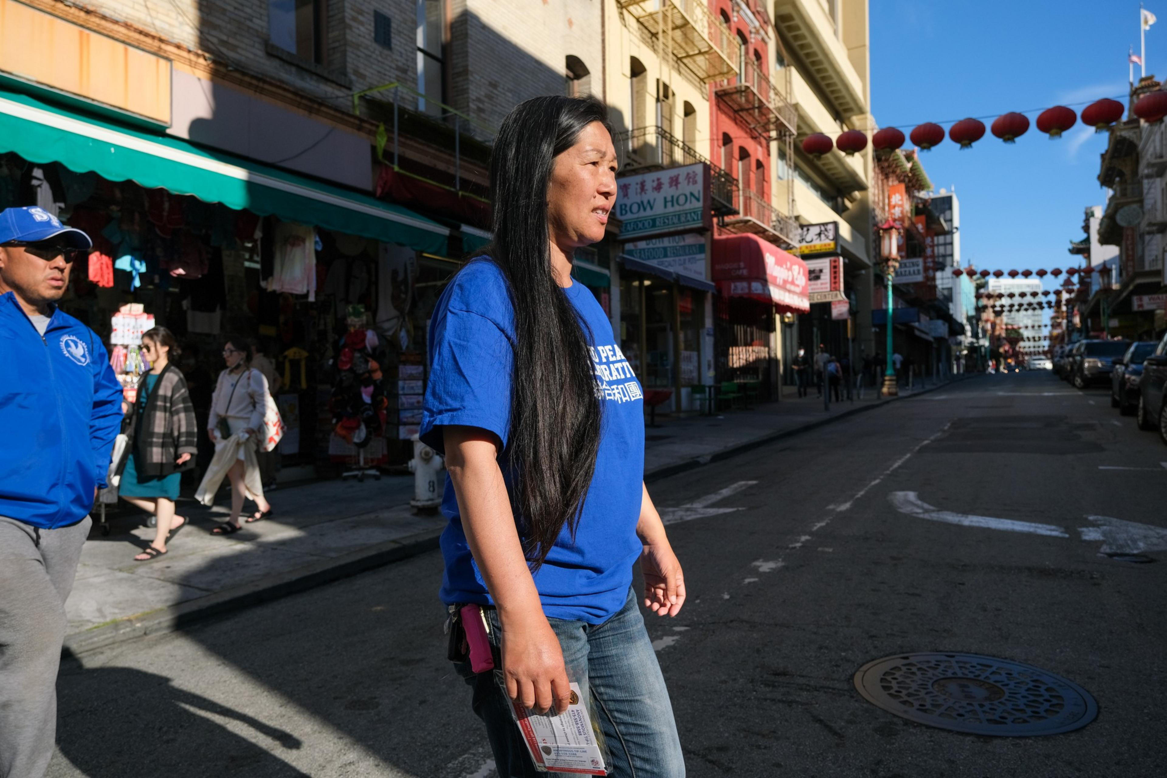 A woman with long black hair and a blue shirt walks across a sunny street lined with shops and hanging red lanterns.