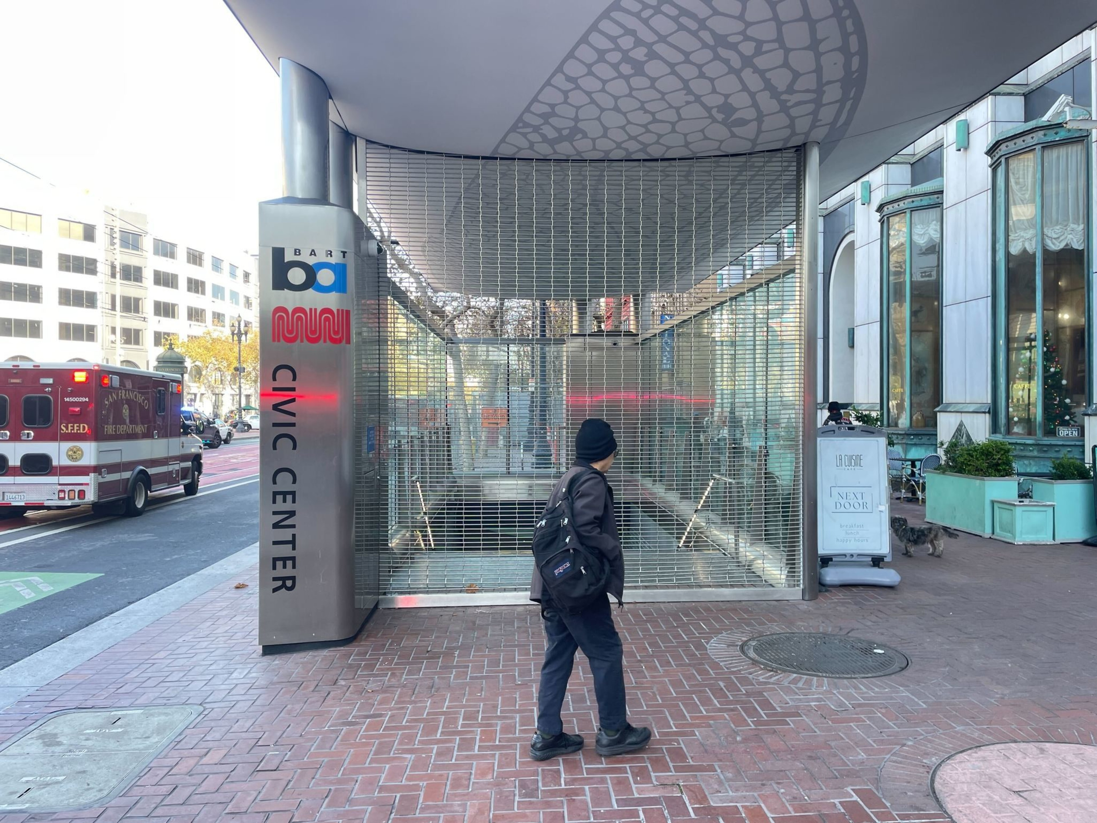 A person with a black backpack and beanie stands in front of a closed Civic Center BART and MUNI subway entrance, with an ambulance on the street nearby.