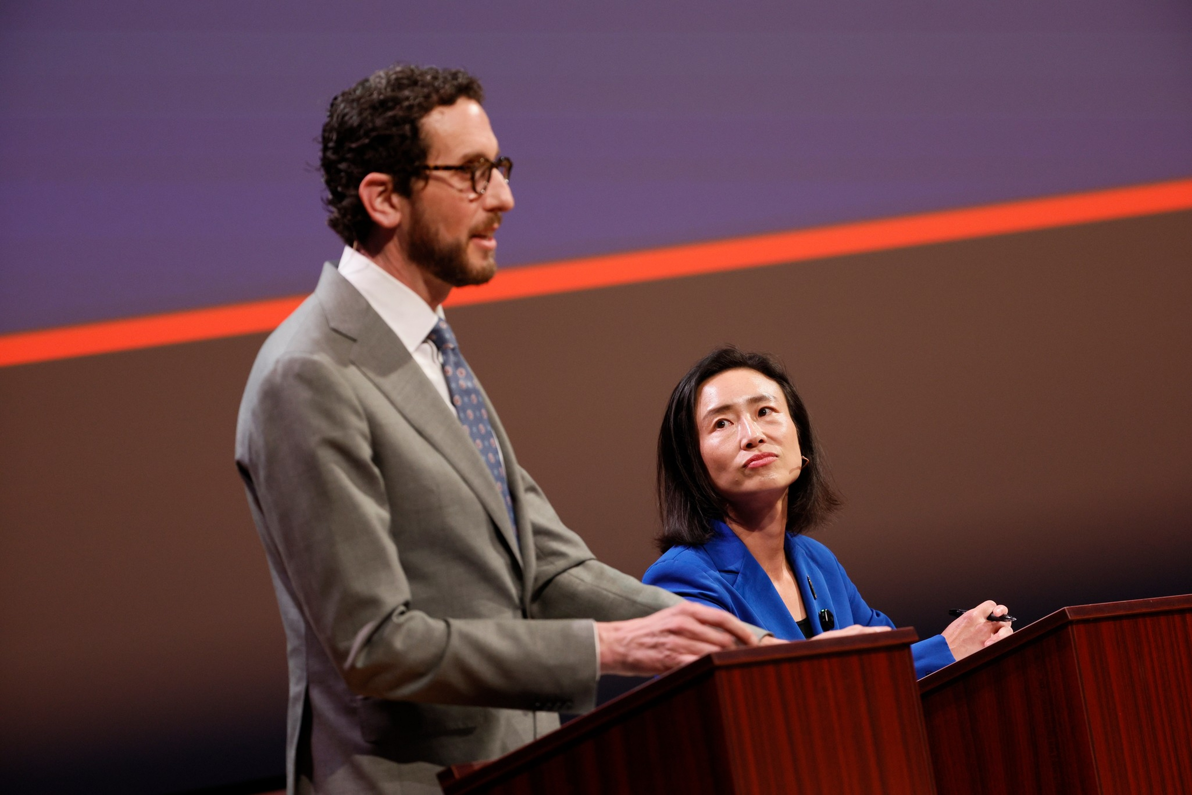 A man in a gray suit speaks at a podium while a woman in a blue blazer listens attentively beside him, both engaged in a formal setting.
