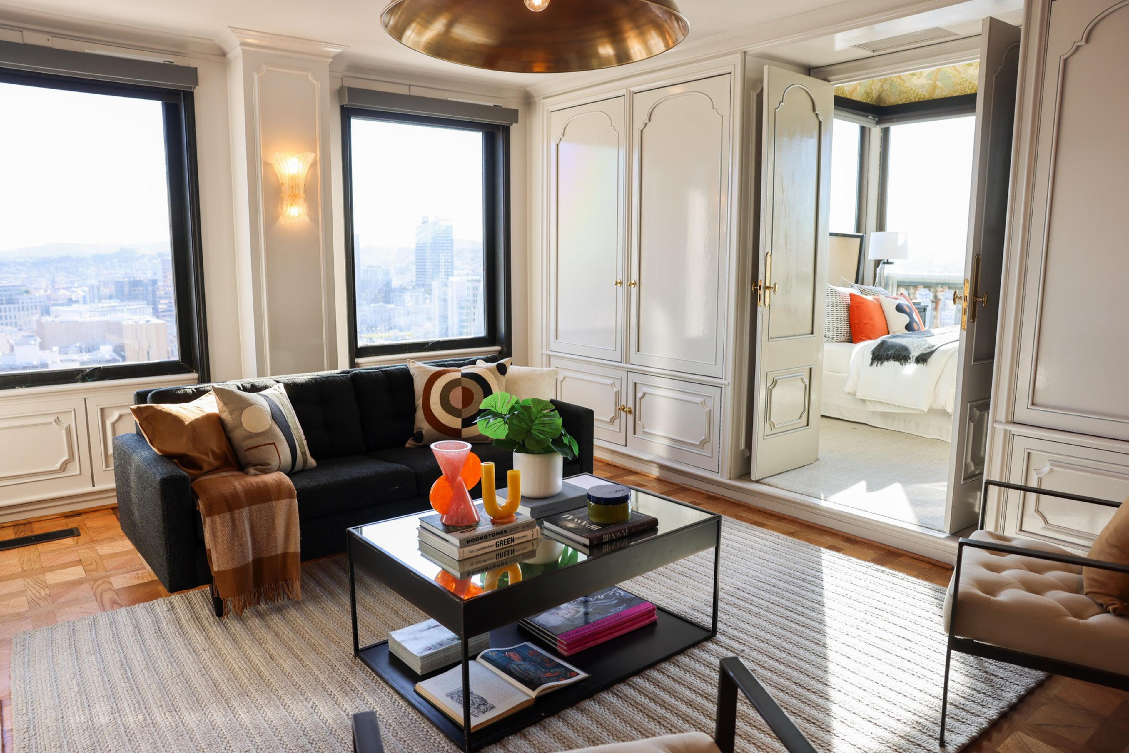 The image shows a modern living room with a black sofa, colorful pillows, a glass coffee table, and large windows. An open door leads to a bright bedroom.