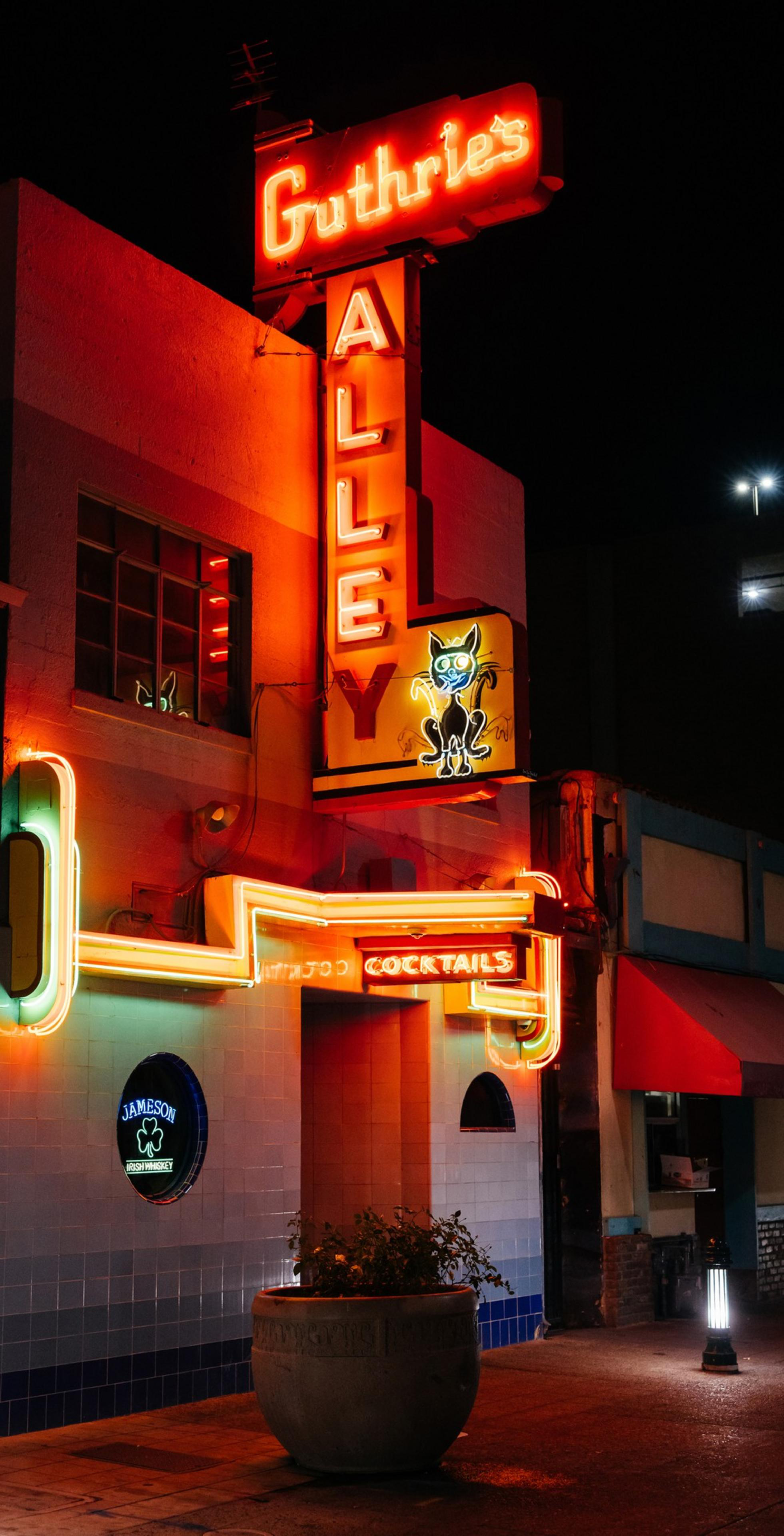 A neon-lit bar sign reads &quot;Guthrie's Alley Cat&quot; with a playful cat illustration. Bright red and yellow lights illuminate the entrance at night.