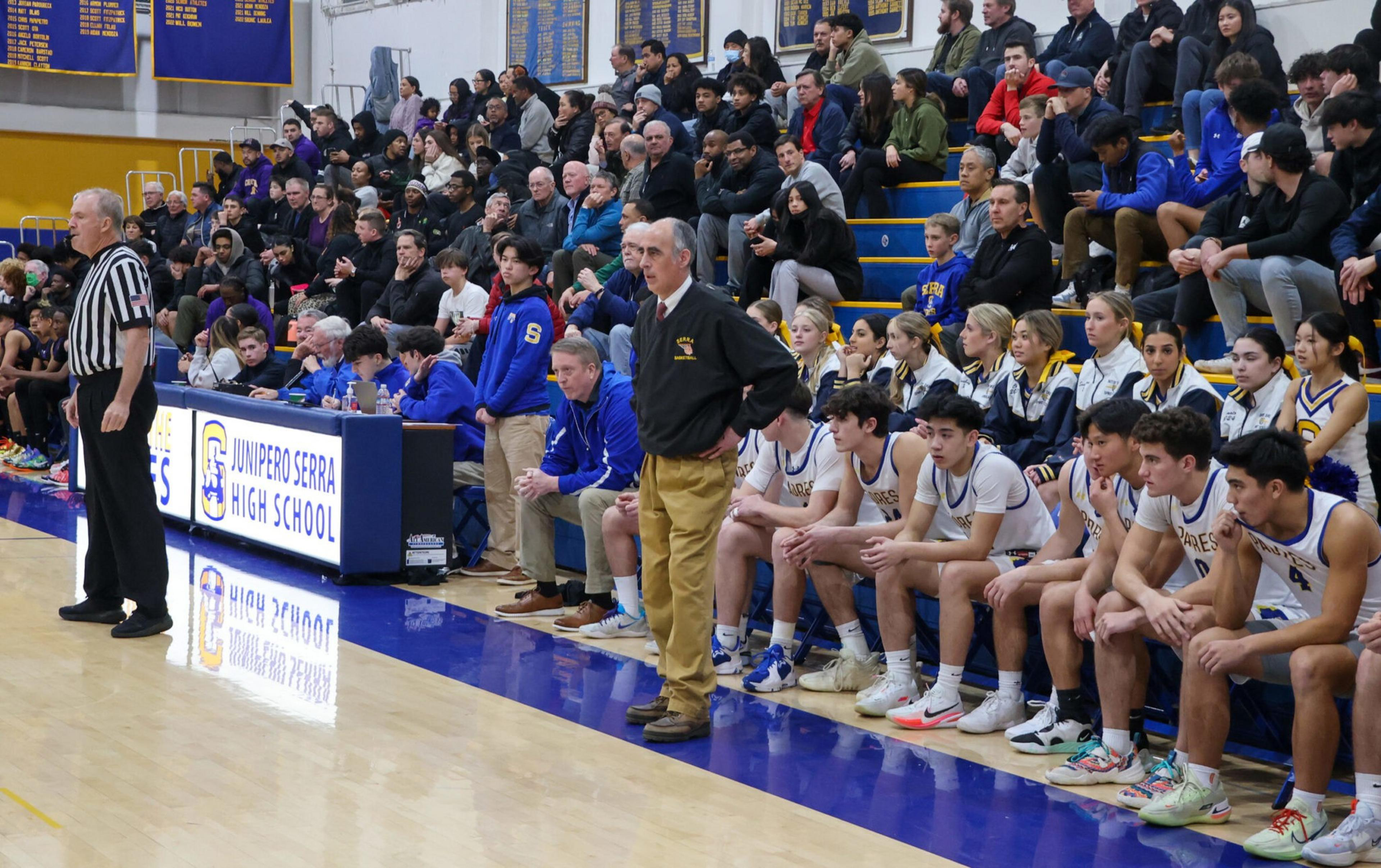 A high school basketball team sits on the bench with coaches standing nearby, while a referee stands on the court and spectators fill the bleachers.