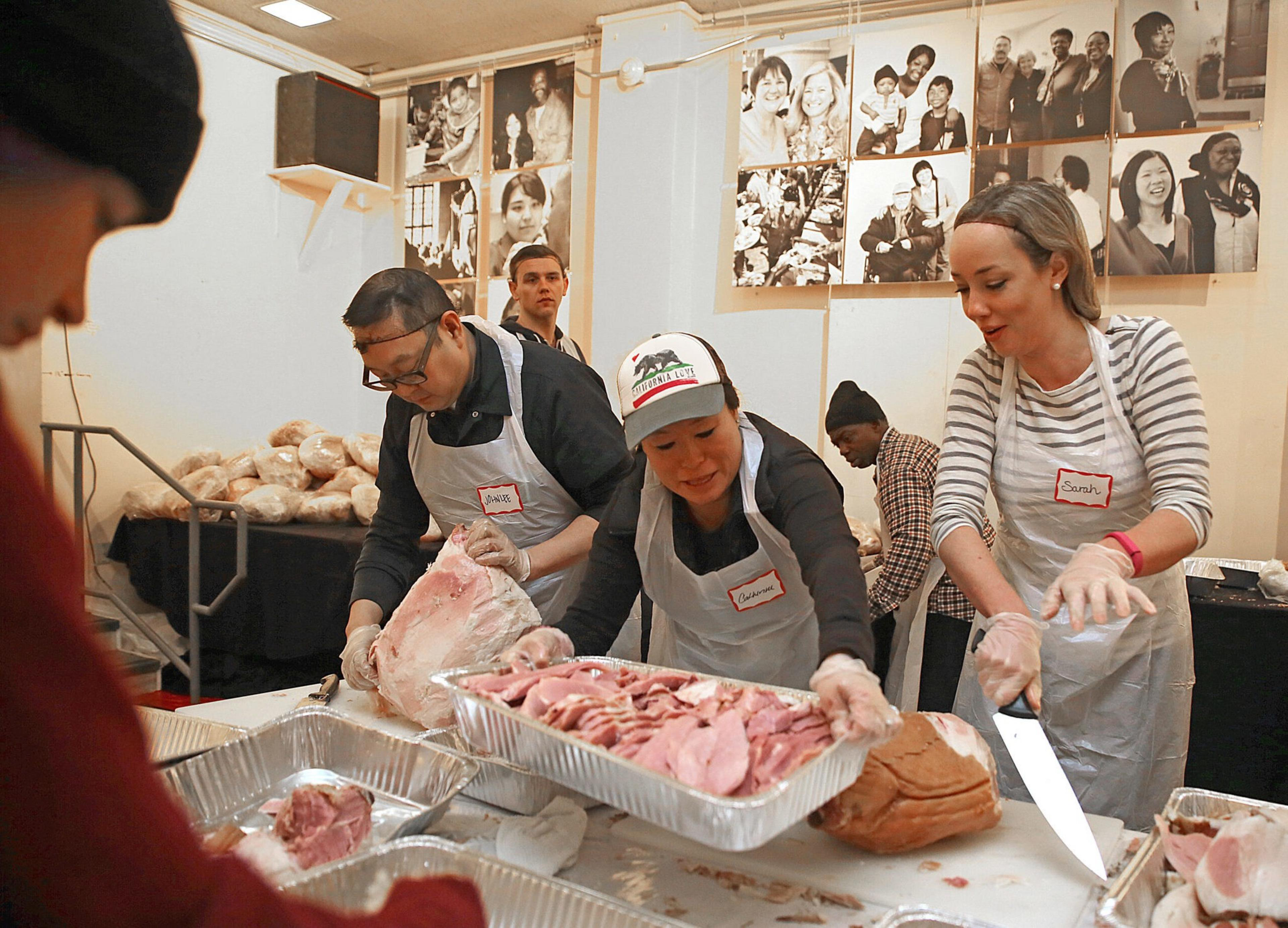 Volunteers wearing aprons and gloves slice and package ham in a community kitchen. Breads are stacked nearby, and black-and-white photos hang on the walls.