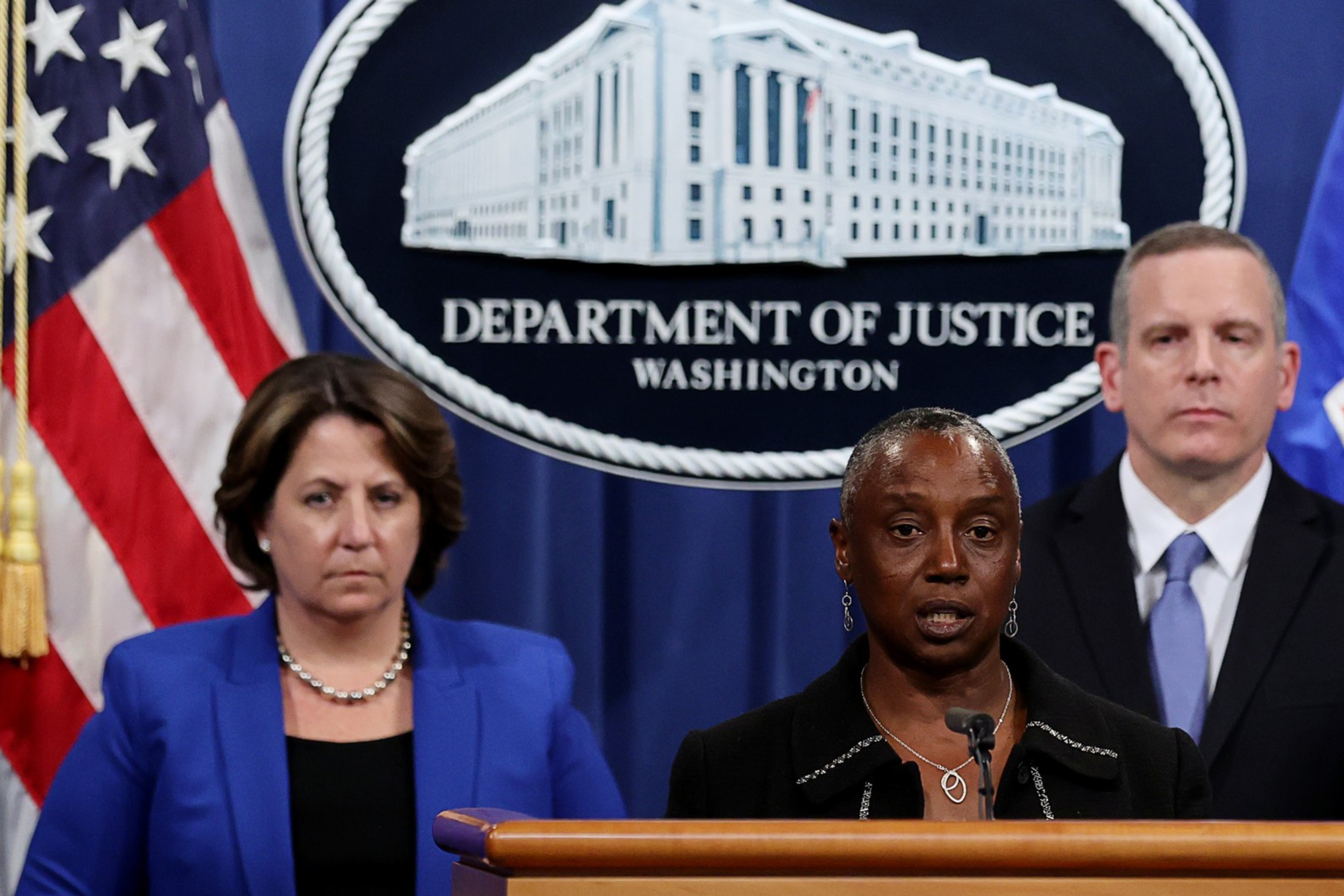 Three officials stand at a podium with the Department of Justice seal. The woman in the center speaks, flanked by a woman in blue and a man in a suit.