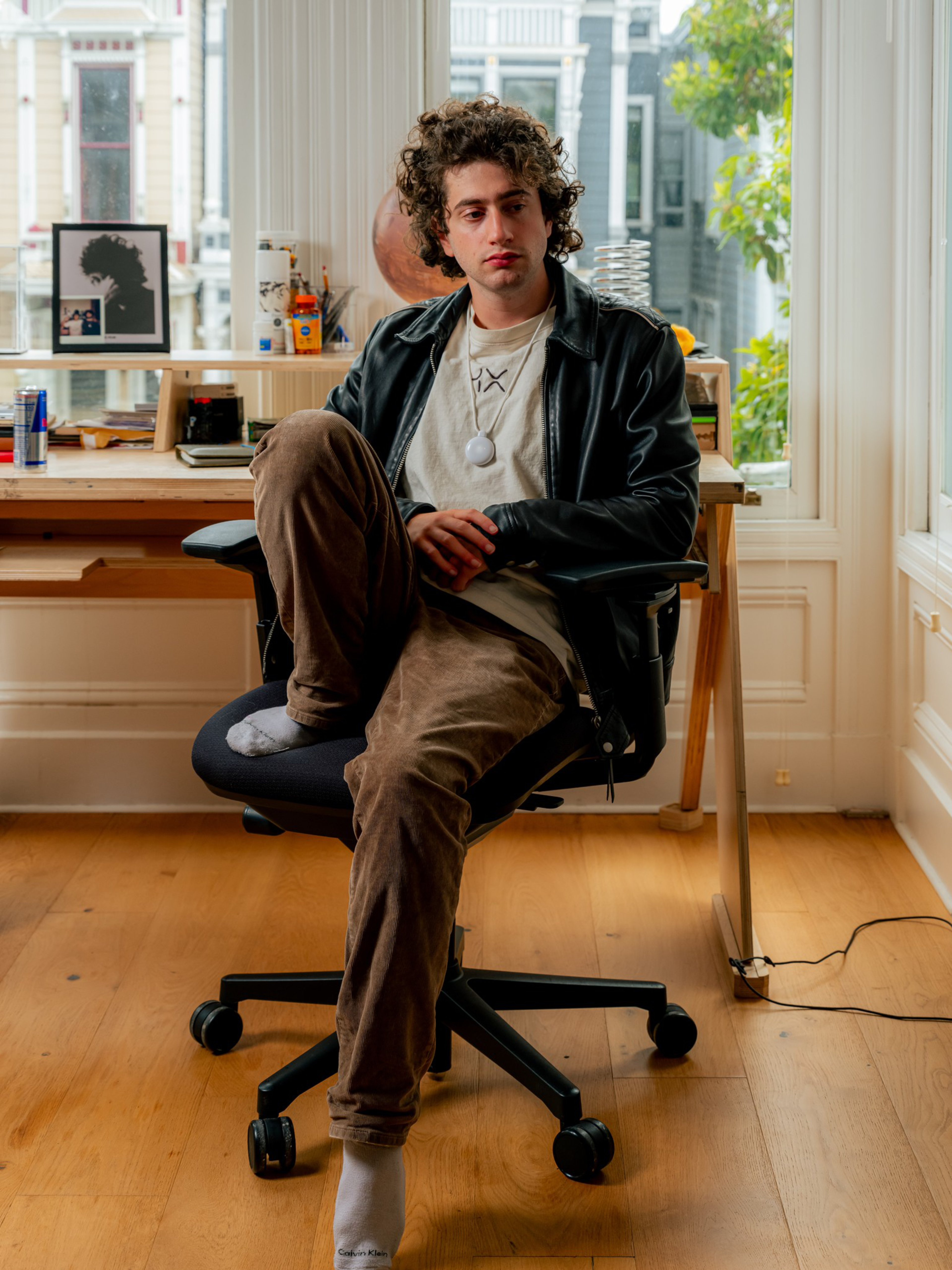A young man with curly hair sits on an office chair with one leg crossed, wearing a leather jacket, beige pants, and white socks.