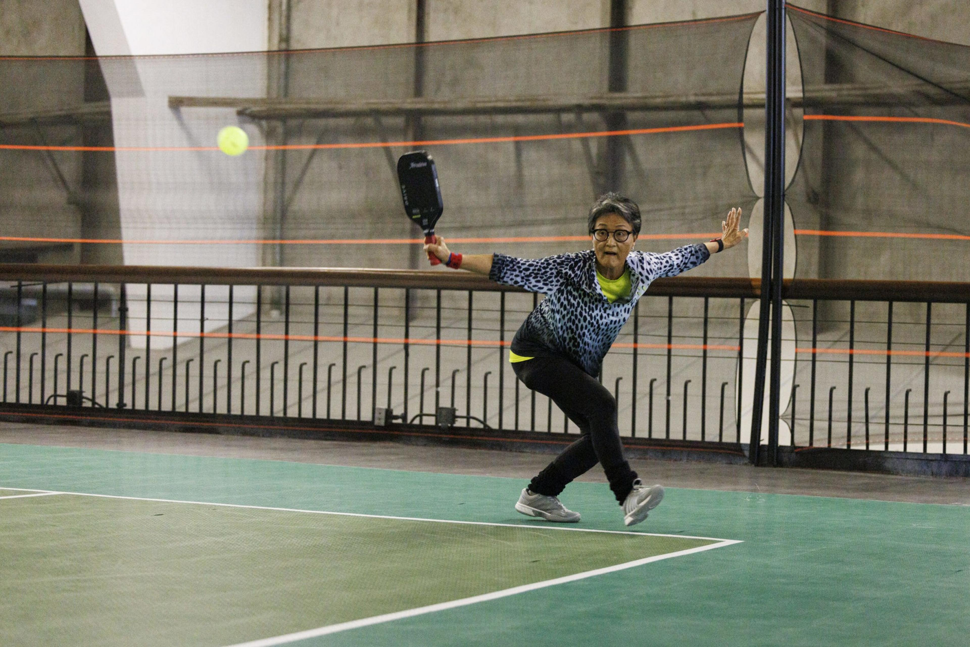 A person in sportswear lunges with a paddle to hit a yellow ball on an indoor pickleball court with a net and a metal railing in the background.