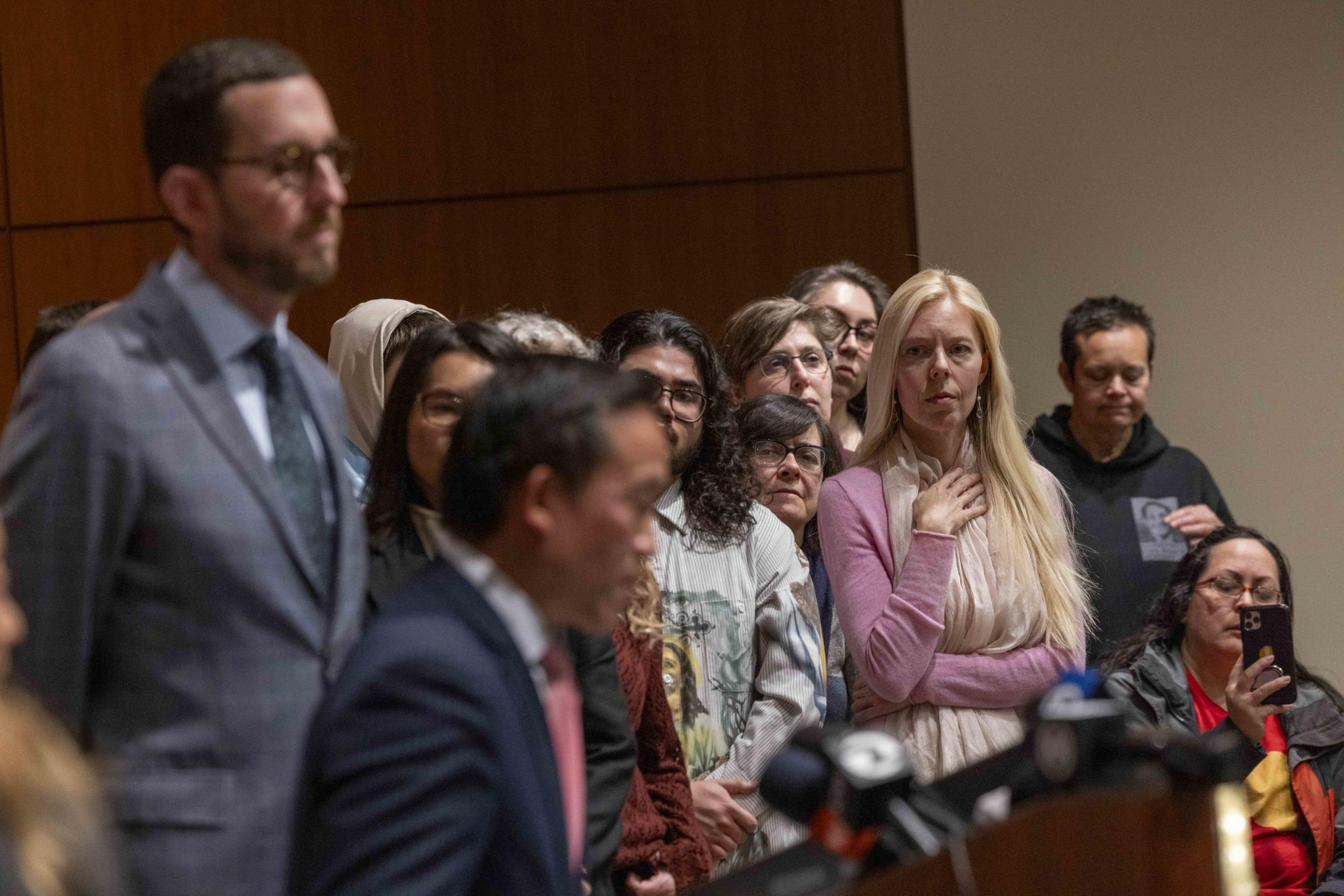 A tall man in a gray suit delivers remarks at a wooden podium flanked by a crowd of people.