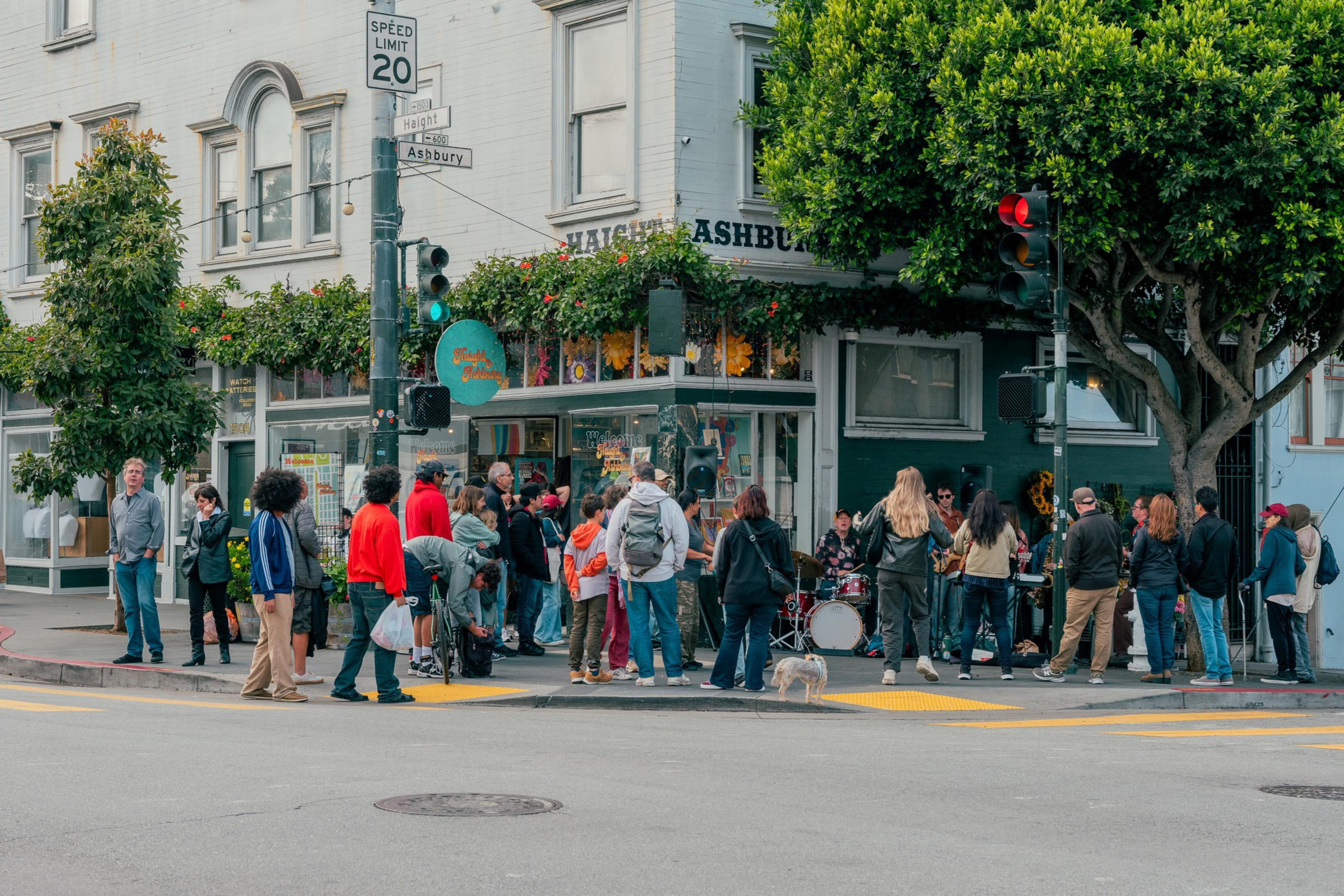 A crowd of people gathers on a street corner under green street signs reading "Haight" and "Ashbury," near a building covered in plants and flowers.