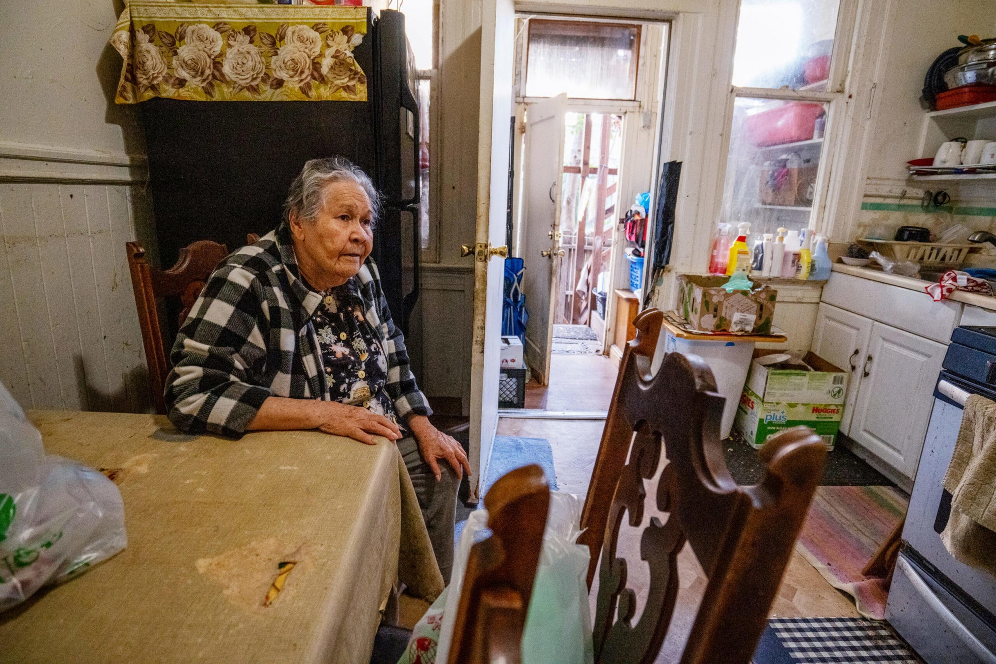 An elderly woman sits at a kitchen table with a plaid shirt, surrounded by wooden chairs, kitchen cabinets, and a doorway leading outside.