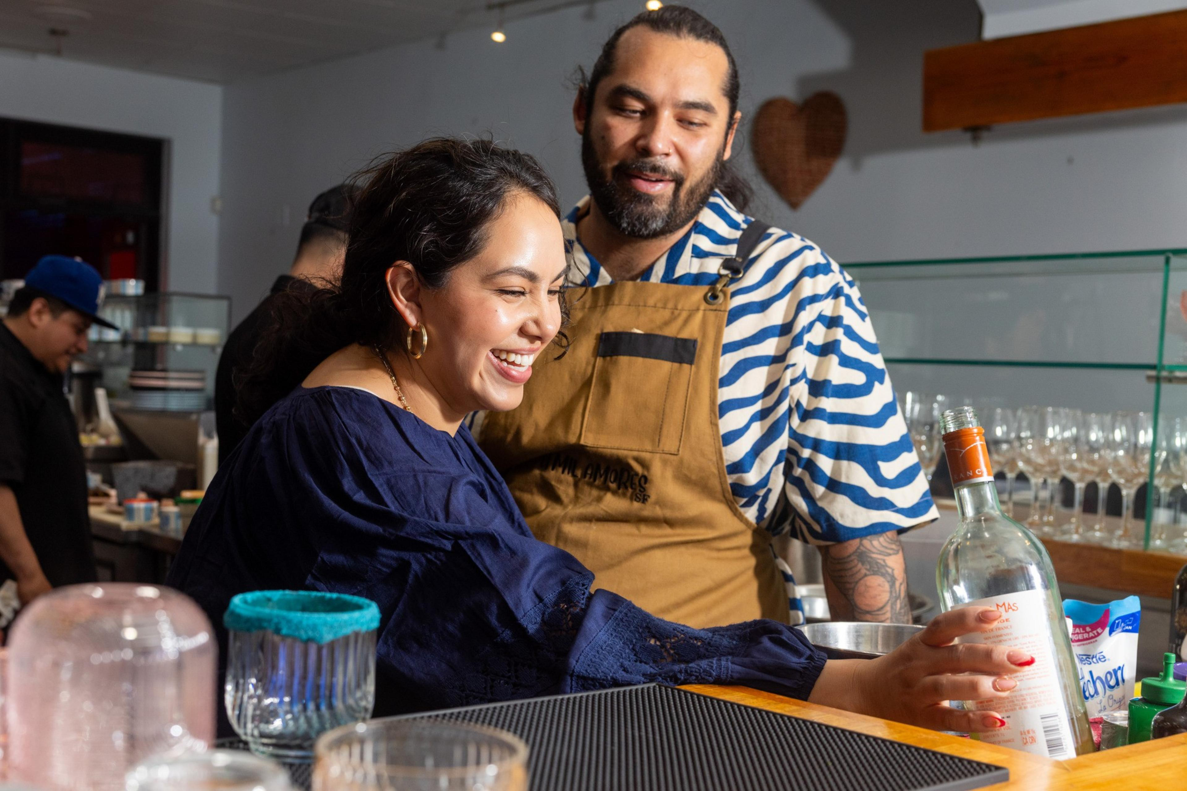 A woman in a blue blouse smiles while holding a bottle, assisted by a man in a striped shirt and apron. They're in a bar setting, with glasses and a counter.