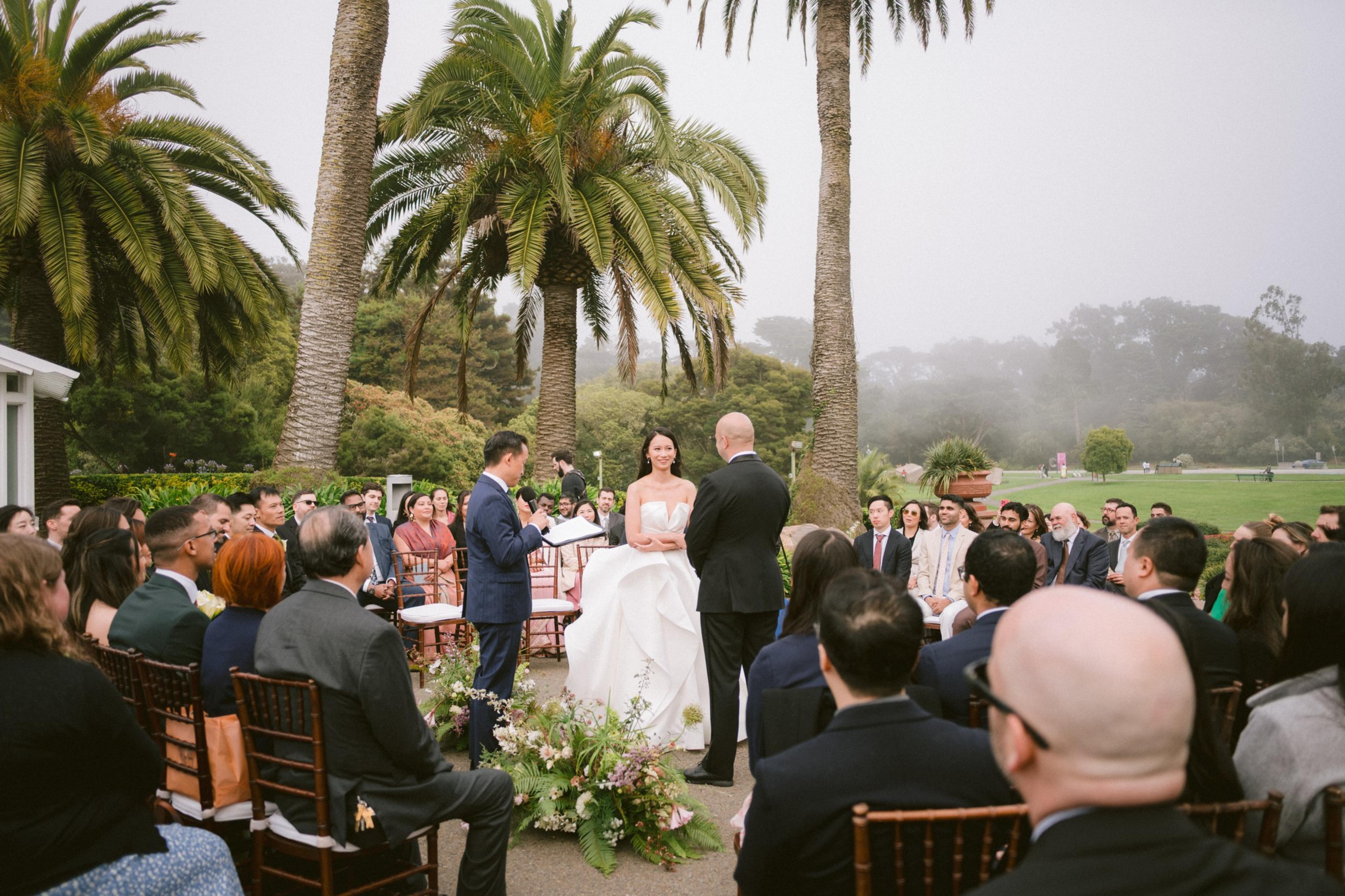 A couple stands before an officiant under tall palm trees, surrounded by seated guests during an outdoor wedding ceremony on a cloudy day.