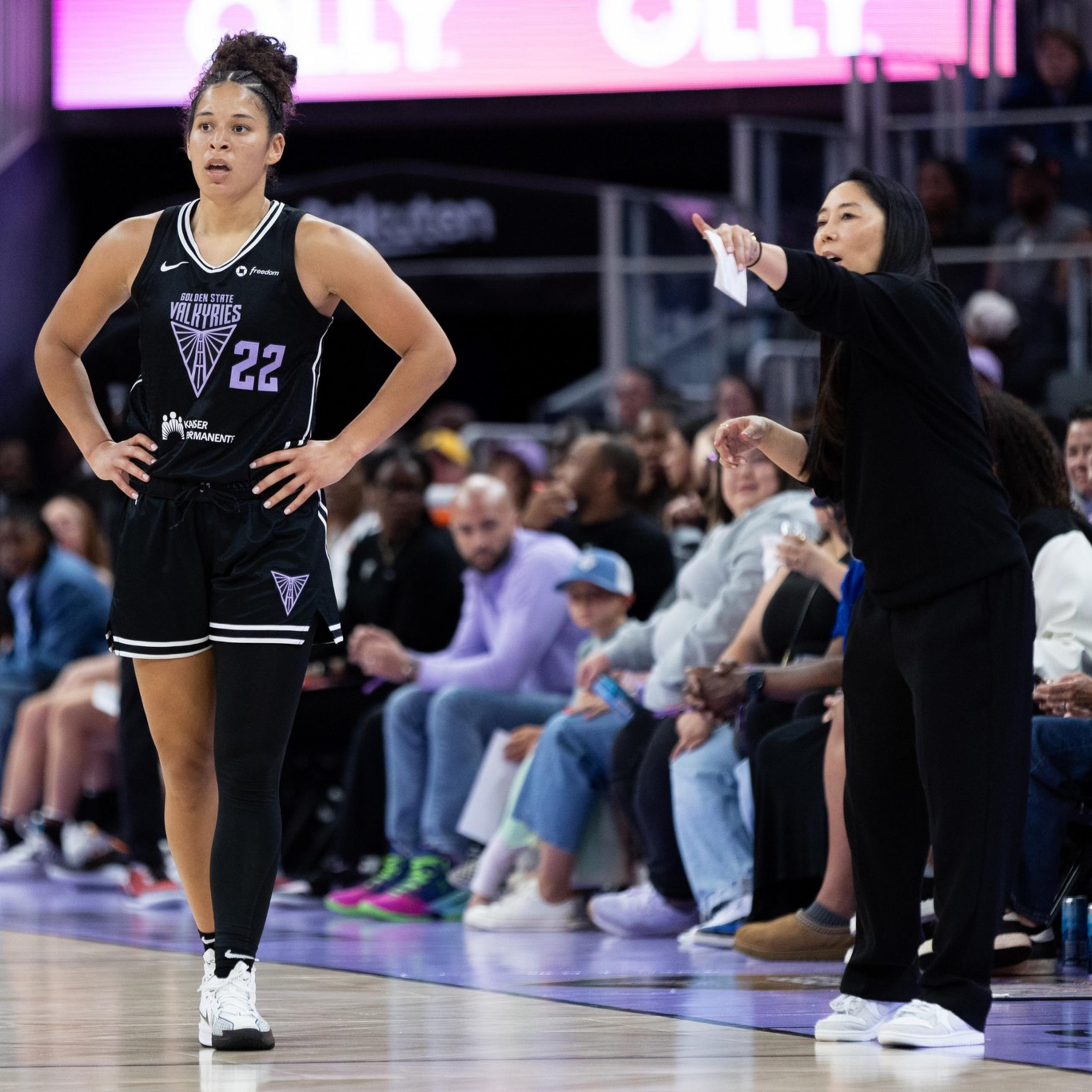 A basketball player stands on the court in a black uniform with "22" on it, while a coach gestures and speaks from the sidelines. Spectators watch in the background.