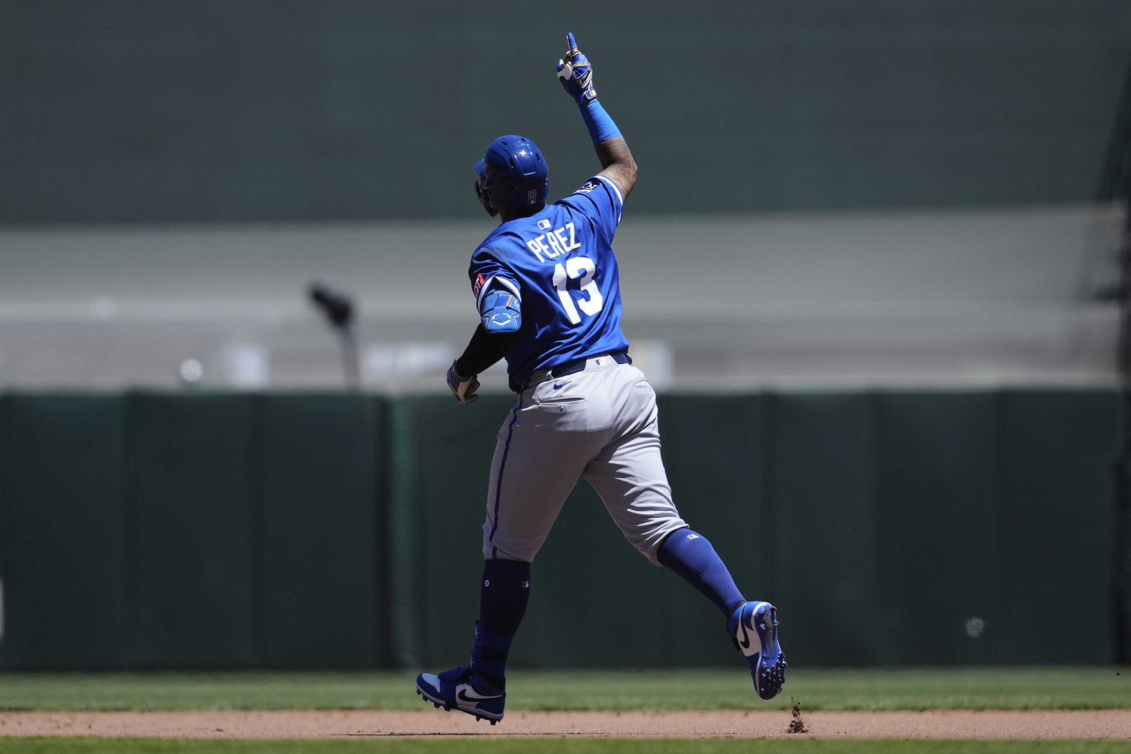 A baseball player wearing a blue uniform with the number 13 raises one arm while running on a field.