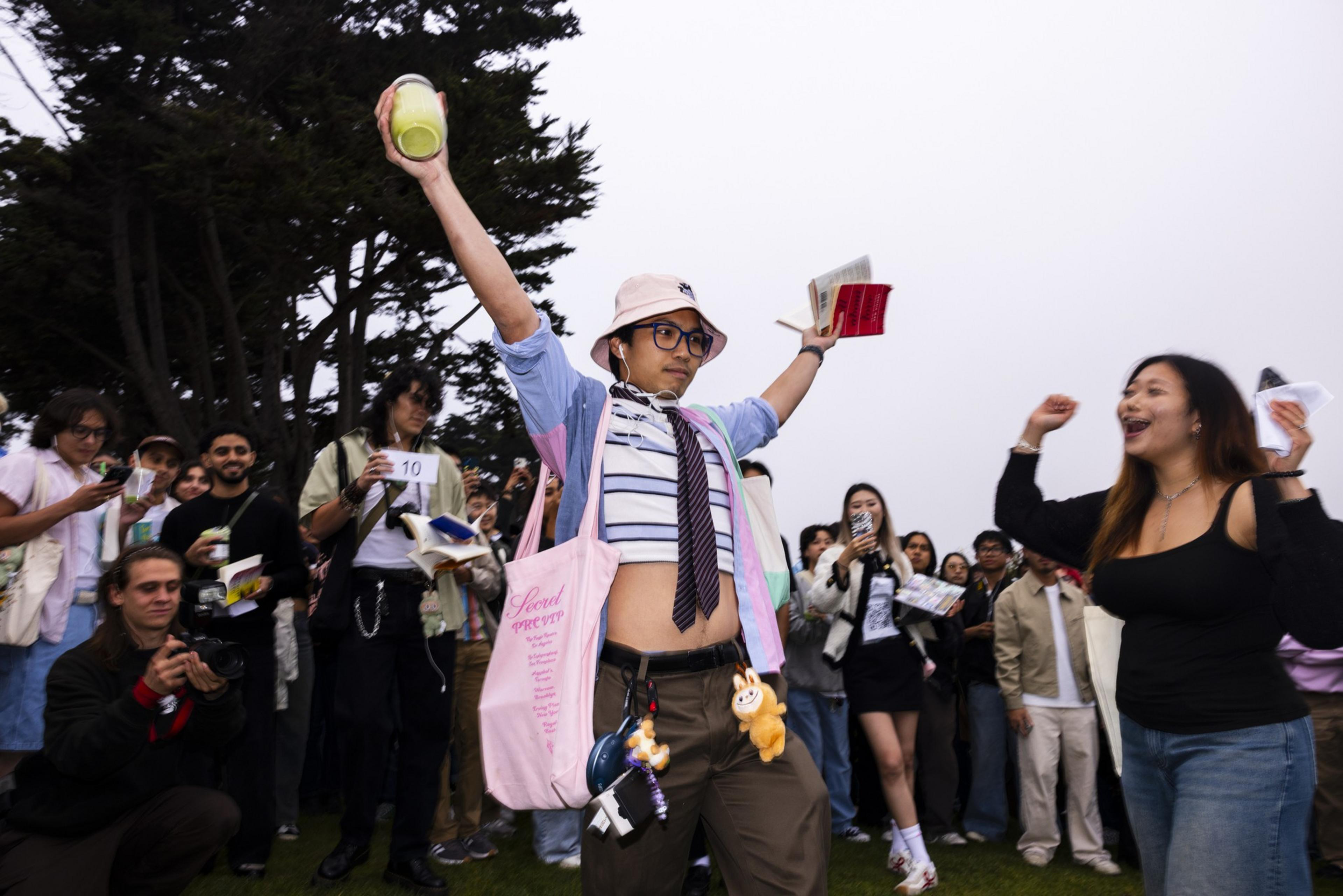 A person wearing a bucket hat, glasses, and a striped crop top raises one arm holding a green drink and the other holding a book, surrounded by cheering people.