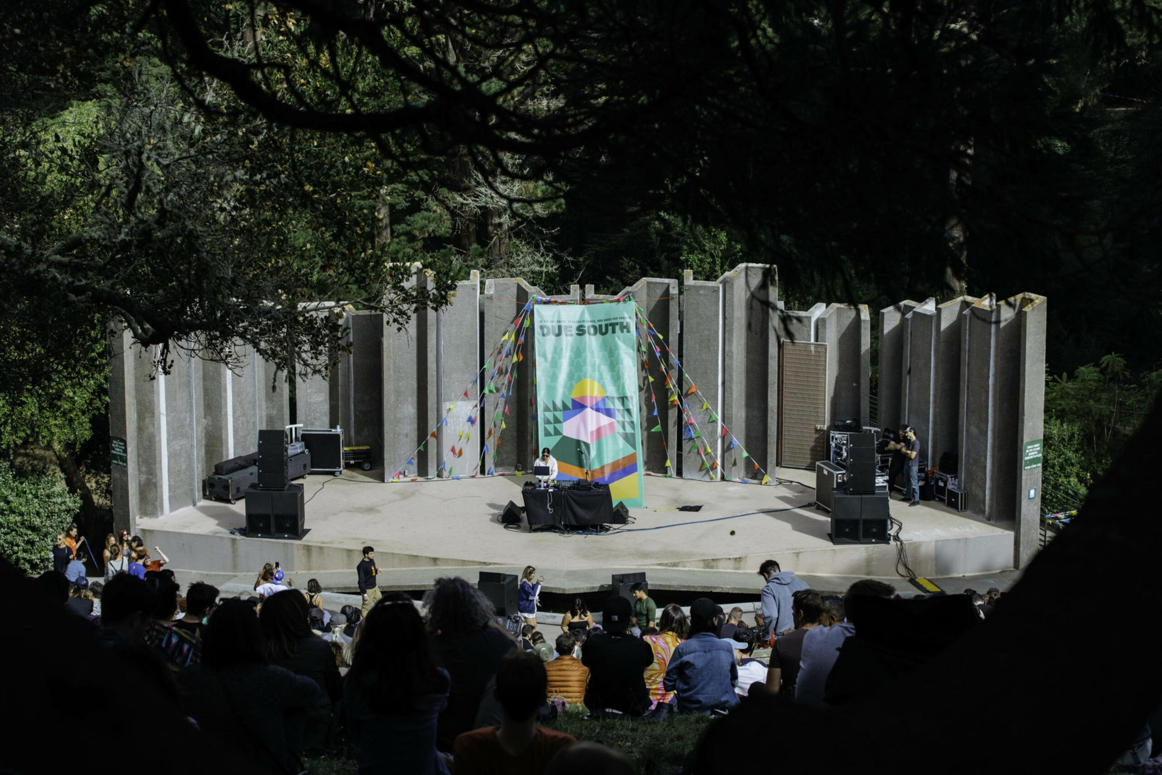 An outdoor stage with a DJ playing under a colorful banner and streamers, surrounded by trees, with a seated audience watching in front.
