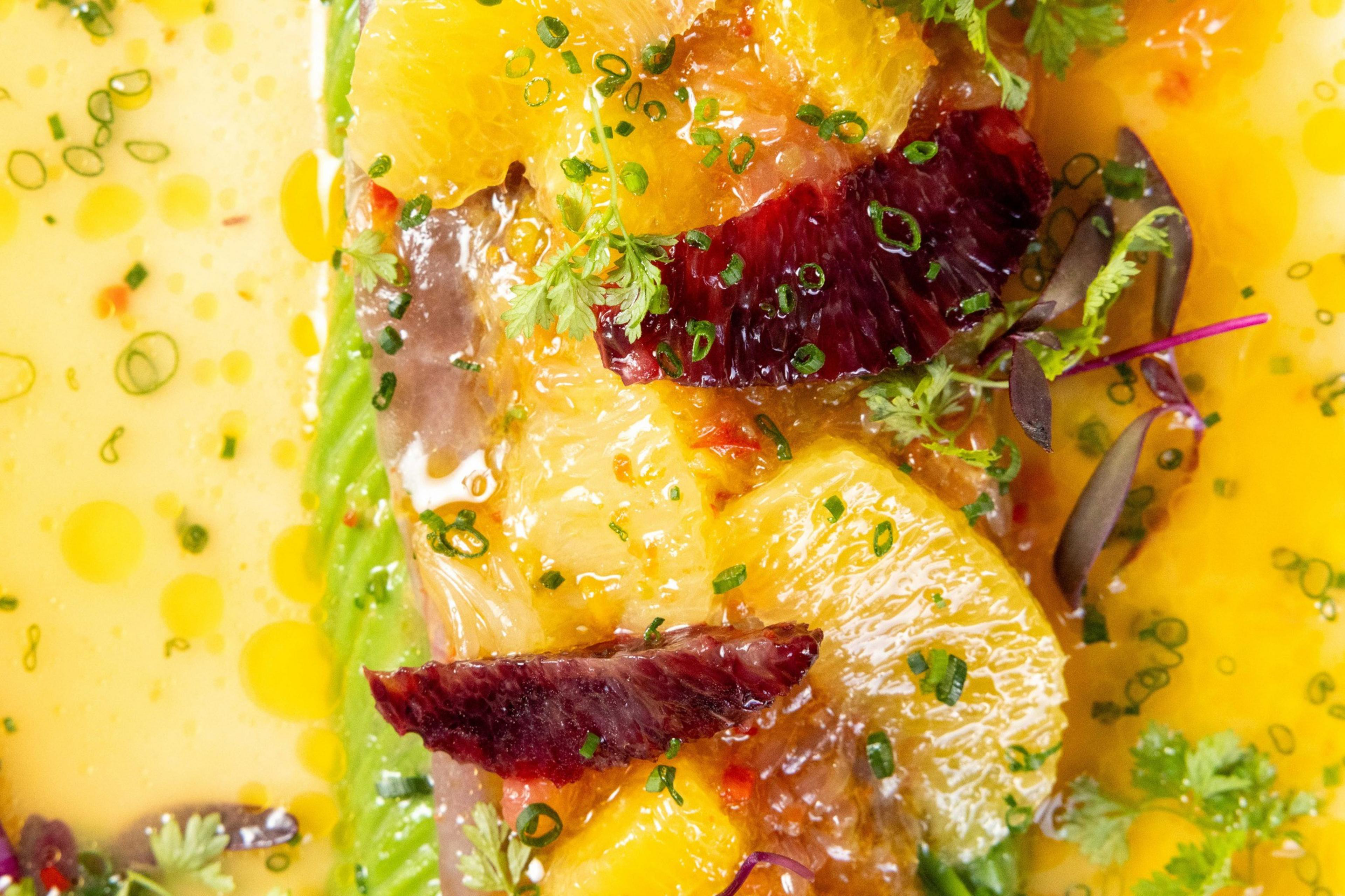 Slices of citrus fruits and raw fish are garnished with herbs and green onions, drizzled with an oily, citrusy sauce.