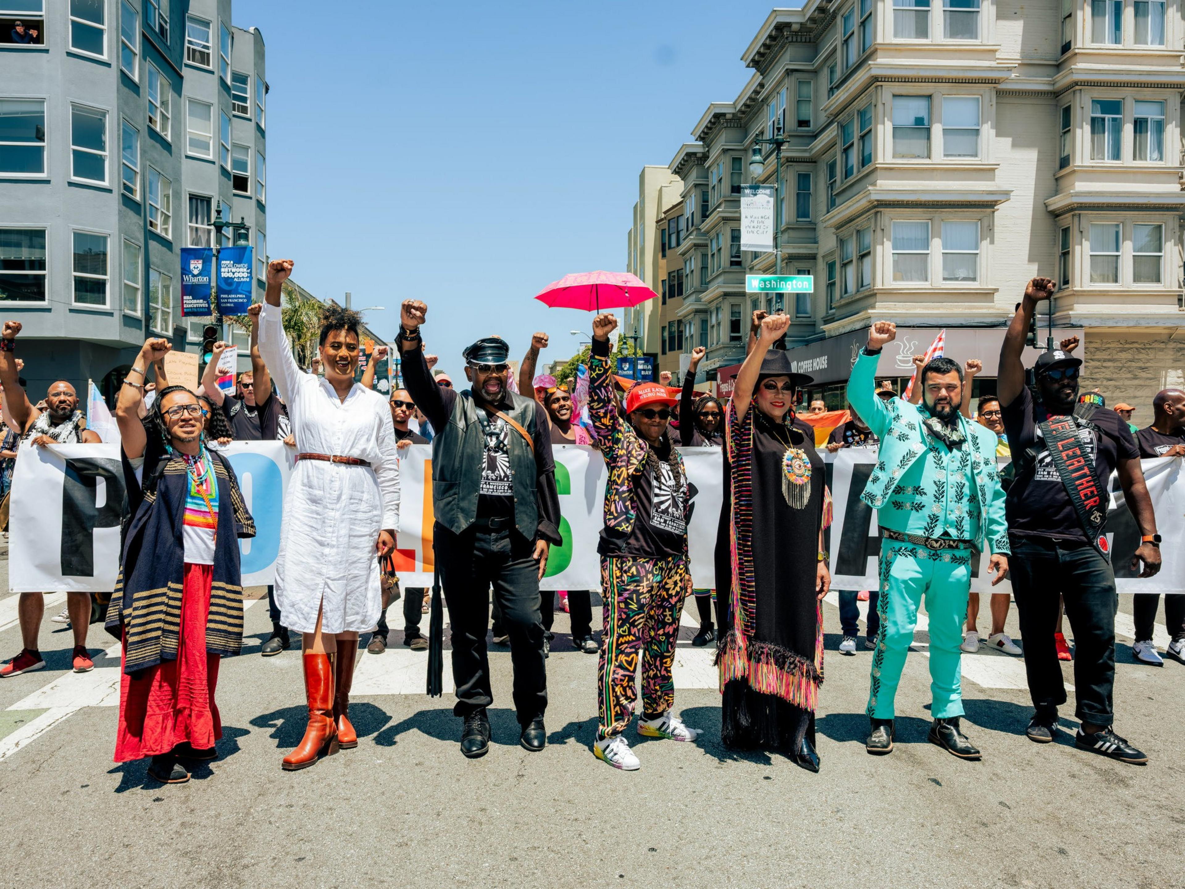A diverse group of people stands on a street, arms raised in unity, holding a banner. The background shows city buildings and the clear blue sky above.