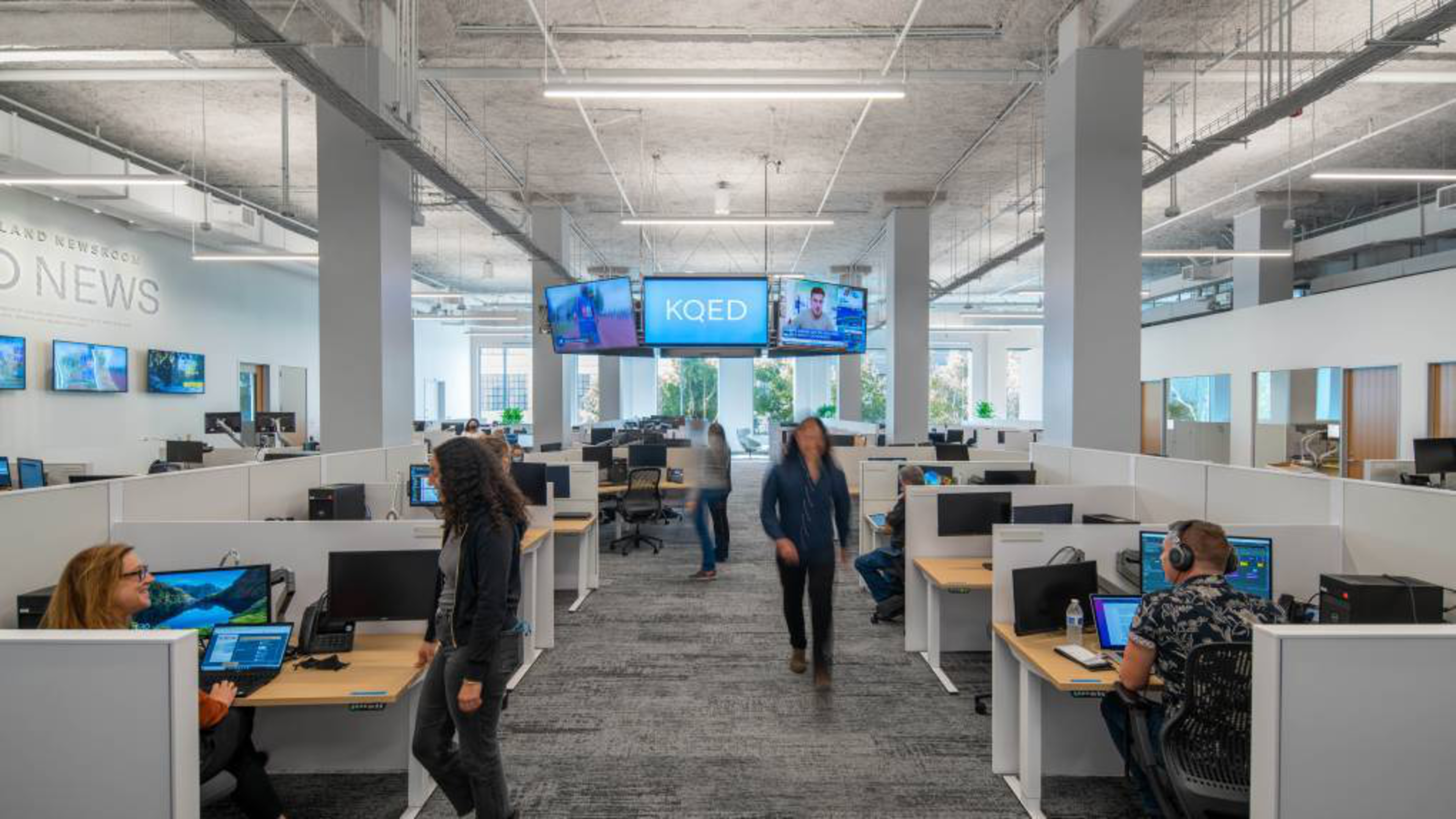 The image shows a modern office with people working at desks, computers, and monitors displaying news. There are large windows and digital screens labeled &quot;KQED.&quot;