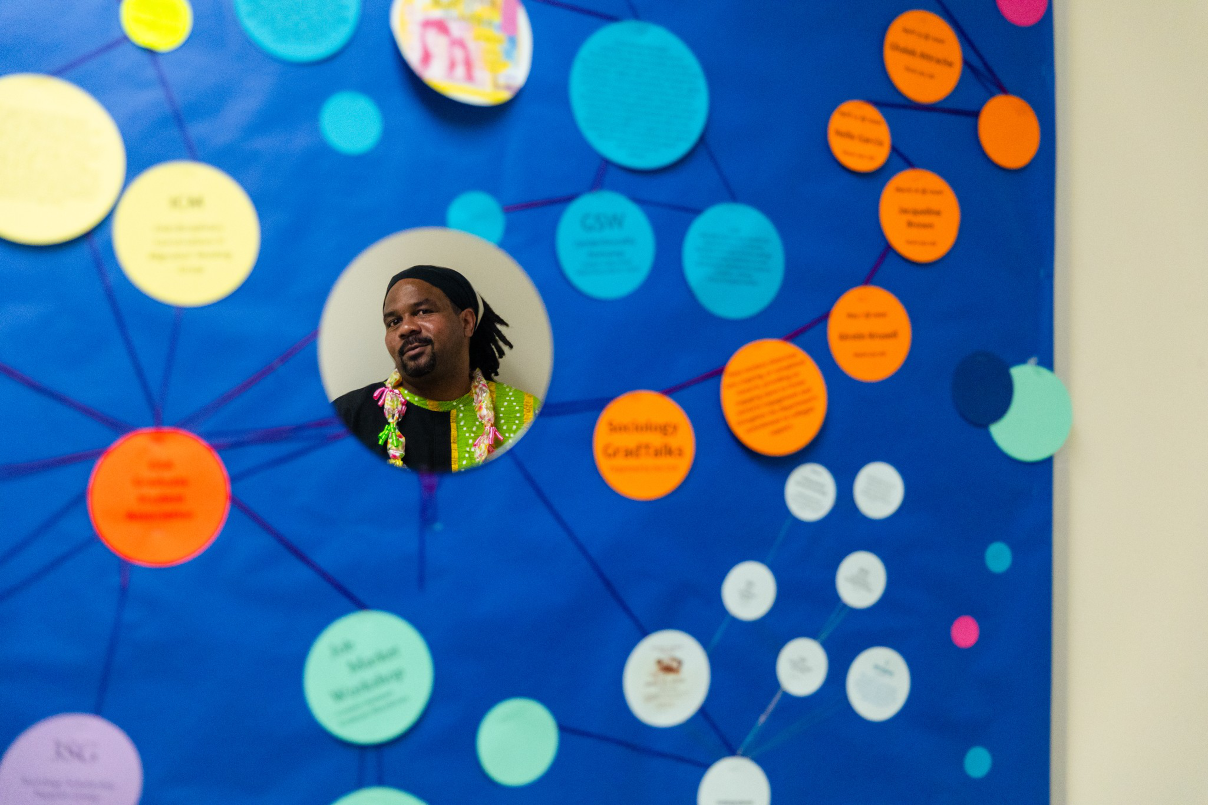 A man with a black headband and colorful shirt is seen through a circular cutout on a blue board filled with multicolored connected circles.