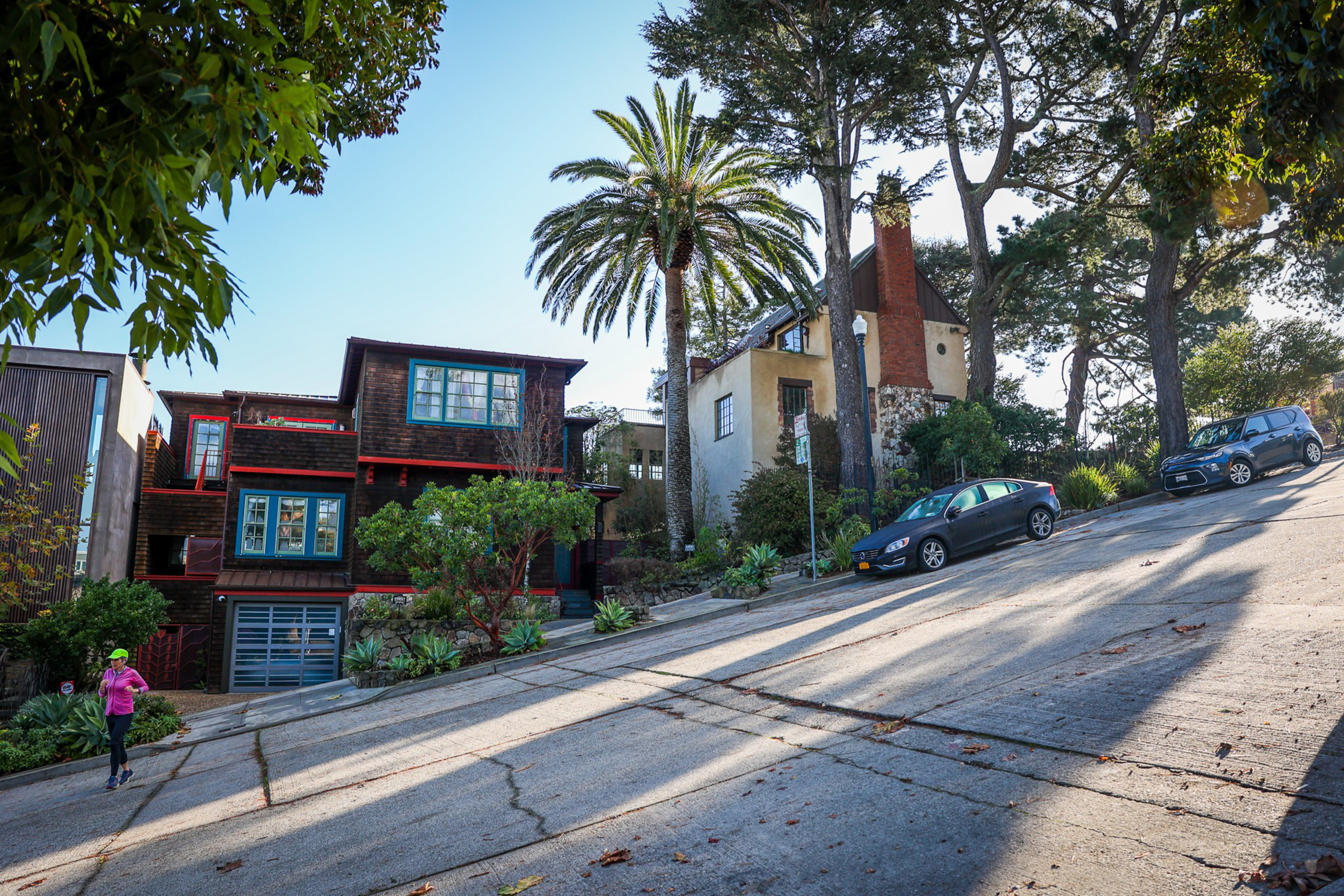 A steep, sloped street with two parked cars, a palm tree, various houses, and a person jogging in bright clothing near the bottom left corner.