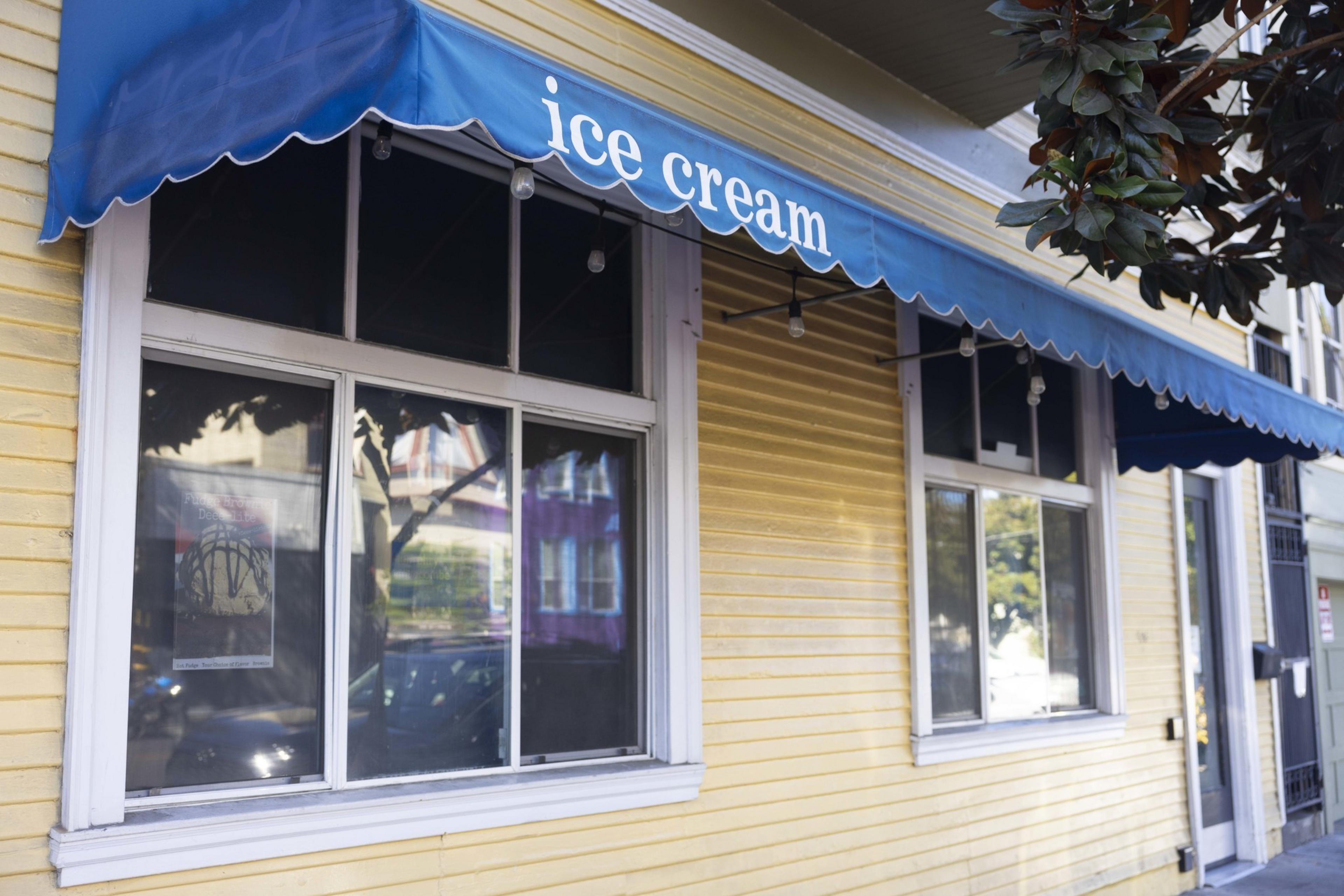 A yellow building with white window frames features a blue awning labeled "ice cream." There are lights strung under the awning, and a tree partially obscures the view.