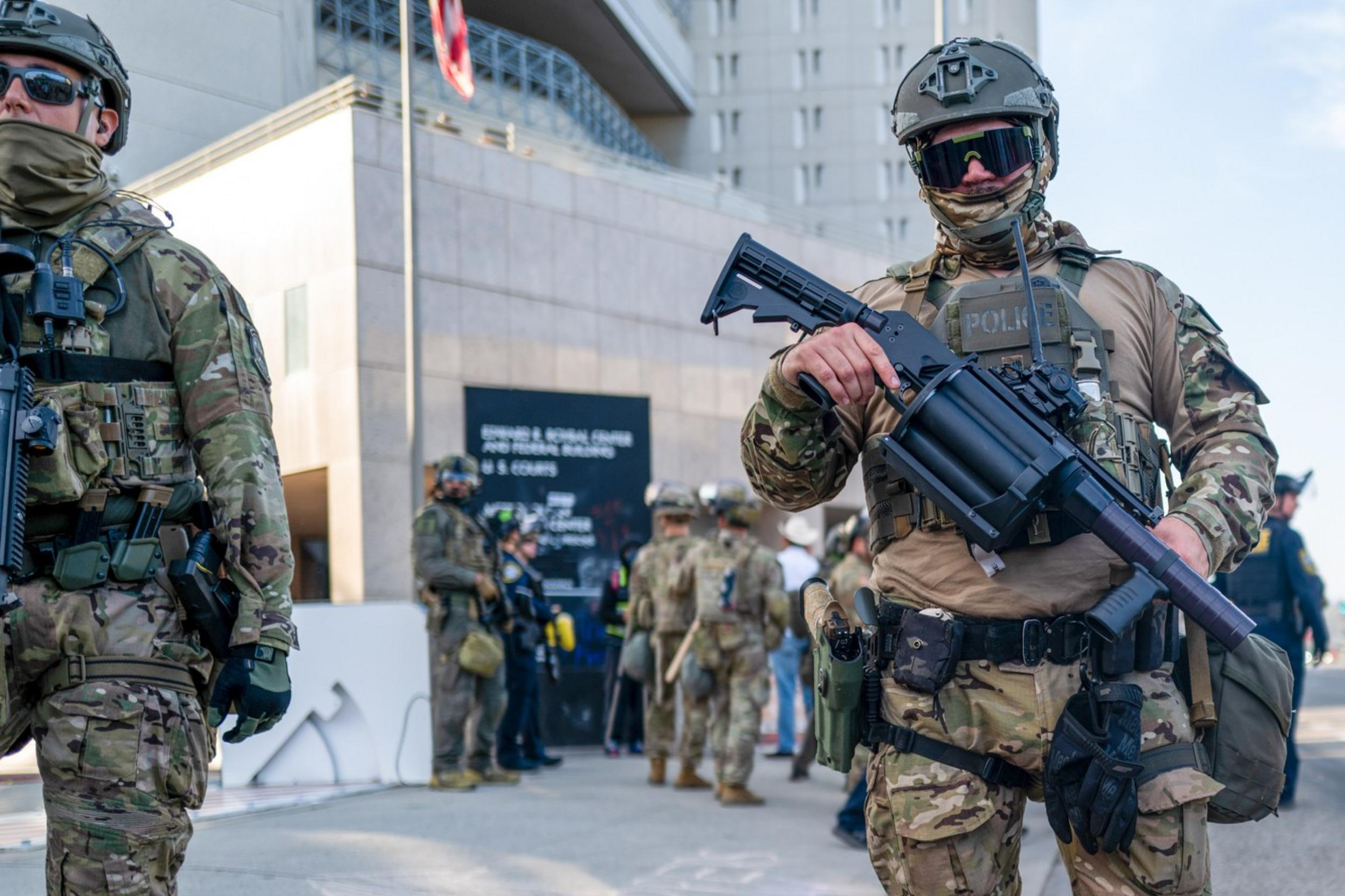 Three armed individuals in tactical gear stand on a street, holding large weapons. They appear to be part of a security detail or law enforcement.